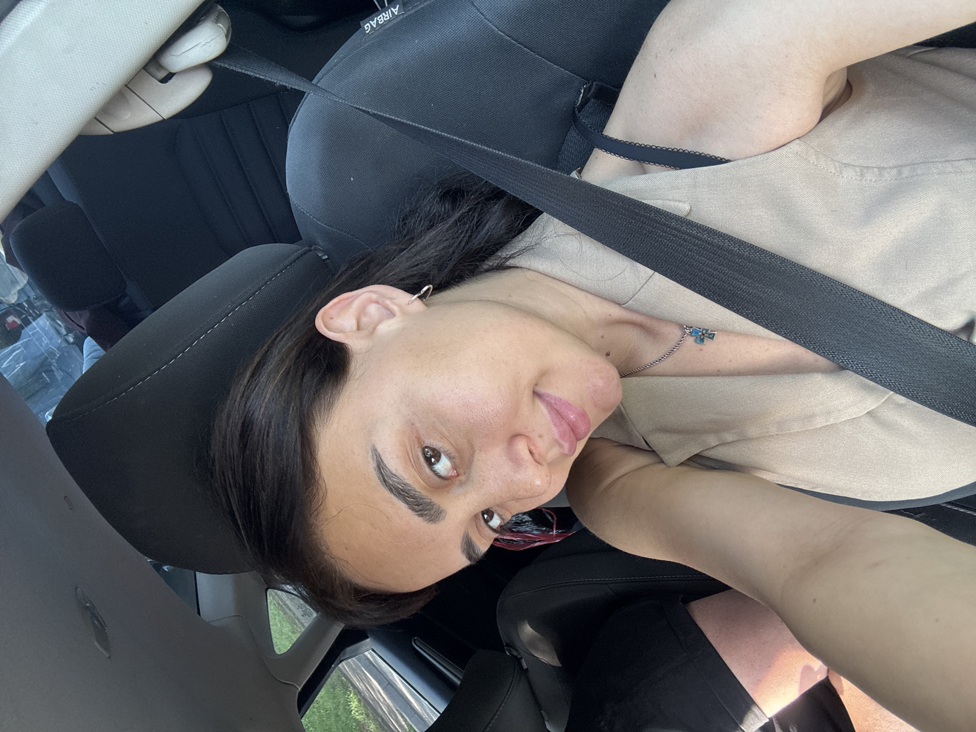 A woman is taking a selfie in the driver's seat of a car. She has dark hair and is wearing a beige vest over a black top. She is also wearing a necklace with a blue pendant and a heart-shaped earring. The car's seatbelt is visible across her chest. The background shows the interior of the car and a glimpse of trees and a road outside the window. The image was taken in Tbilisi, Georgia.