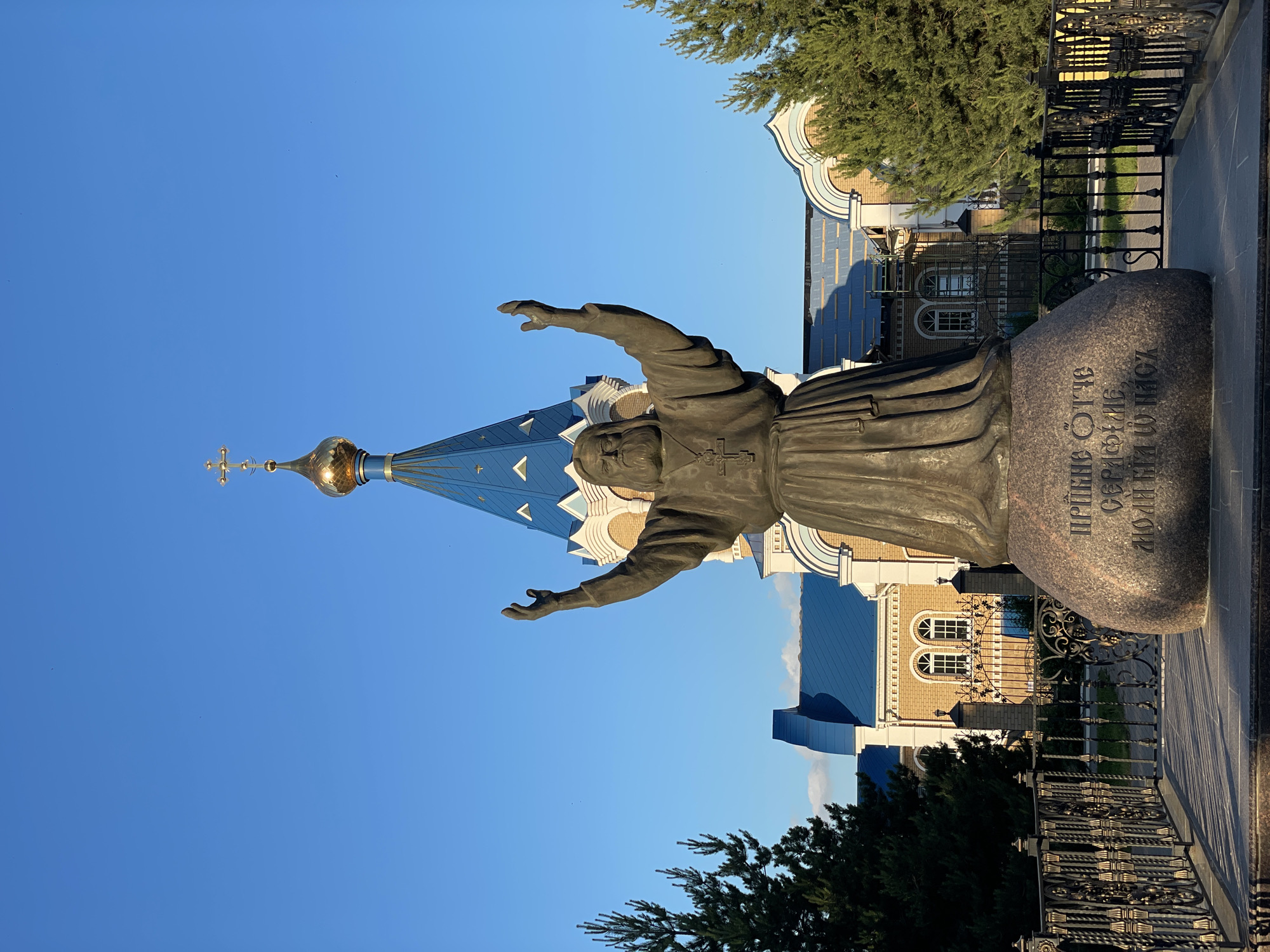 The image shows a bronze statue of a man with a beard, wearing a robe and a cross necklace, with his arms raised. The statue is situated on a large rock inscribed with Russian text. Behind the statue, there is a building with a blue, multi-faceted roof and a golden dome topped with a cross. The building appears to be a church or monastery. The scene is set against a clear blue sky. The statue and building are surrounded by greenery and a decorative metal fence. The inscription on the rock reads "ПРИДИ ОТЧЕ СЕРАФИМЕ, МОЛИТВУ И НАШ", which translates to "Come, Father Seraphim, and our prayer". This inscription, along with the building's architecture, suggests a religious context. The statue is a monument to Saint Seraphim of Sarov, a prominent Russian Orthodox mystic. The building is likely the Seraphim-Diveyevo Monastery in Izhevsk, Russia, or a related religious site dedicated to him.