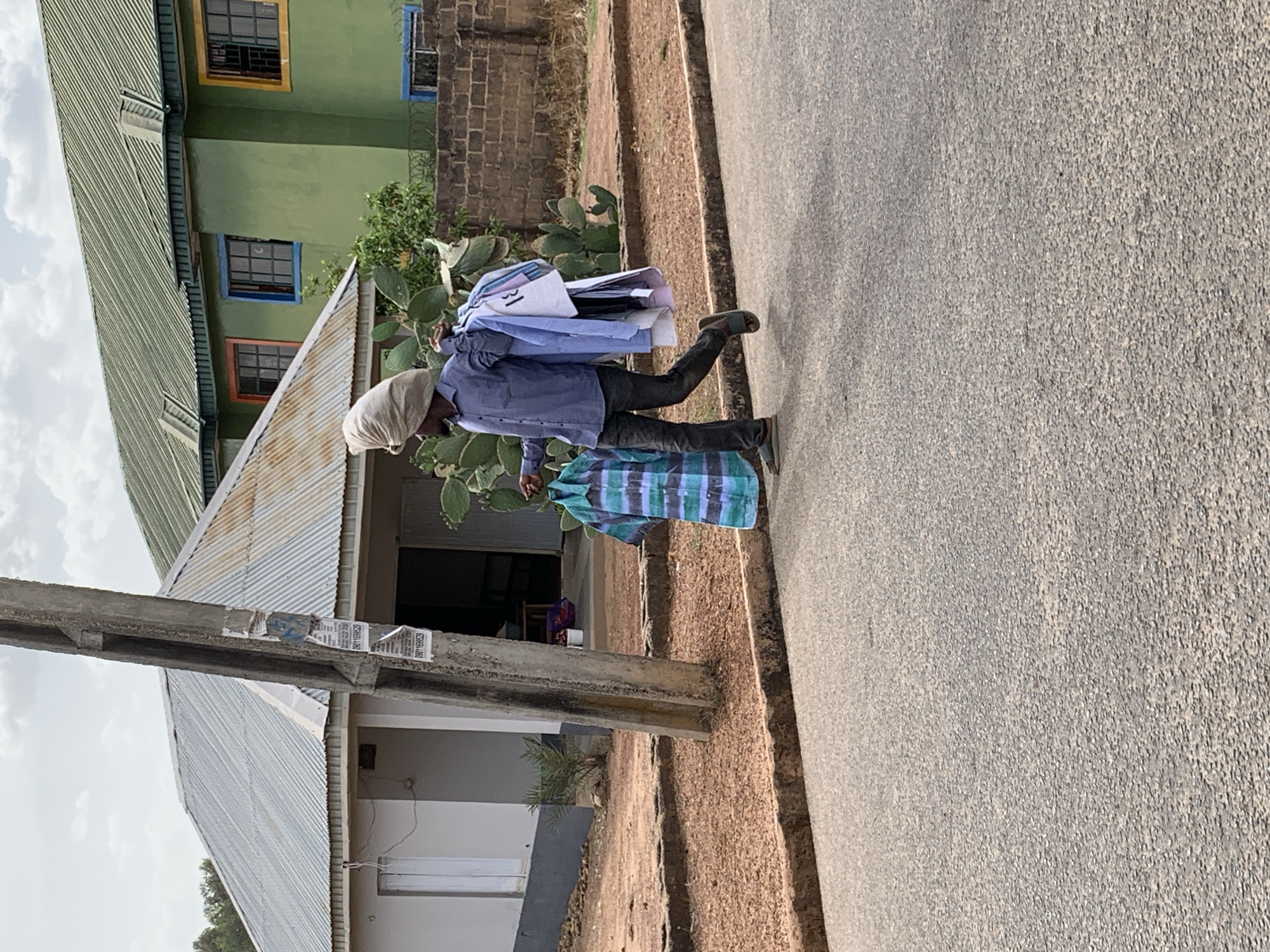 A man walks along a gray asphalt road in what is likely a residential area. The man is wearing a light blue button-down shirt, dark pants, and dark sandals. He also has a light-colored head covering. He carries a stack of clothing over his shoulder. The clothing appears to be a mix of light-colored shirts. On his left side, hanging down to his knees, is a striped teal and white plaid cloth.
Behind him is a building with a corrugated metal roof. A utility pole stands beside the building, with pieces of paper stuck to it. Behind the metal roof building is a larger green structure with multiple windows. The scene is likely located in Jos, Nigeria, based on the style of the buildings and the appearance of the man.