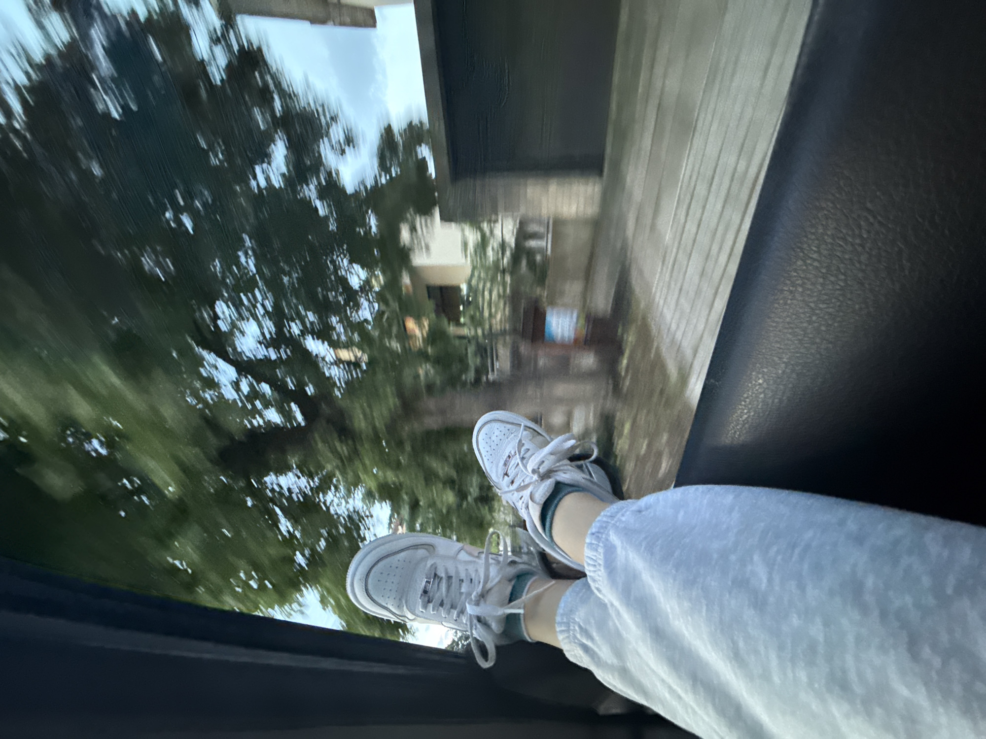 The photograph shows a view from inside a moving vehicle, likely a car. A pair of white sneakers and light-colored sweatpants are visible in the foreground, positioned near the open window, suggesting someone is dangling their legs out of the vehicle. Outside the window, the background is blurred, but identifiable as an urban area with buildings and trees. The location appears to be Burgas, Bulgaria, based on the style of architecture, and landscaping visible through the window. The lighting conditions are dim, possibly indicating dusk or overcast weather. The vehicle's interior has dark trim, visible around the window frame.