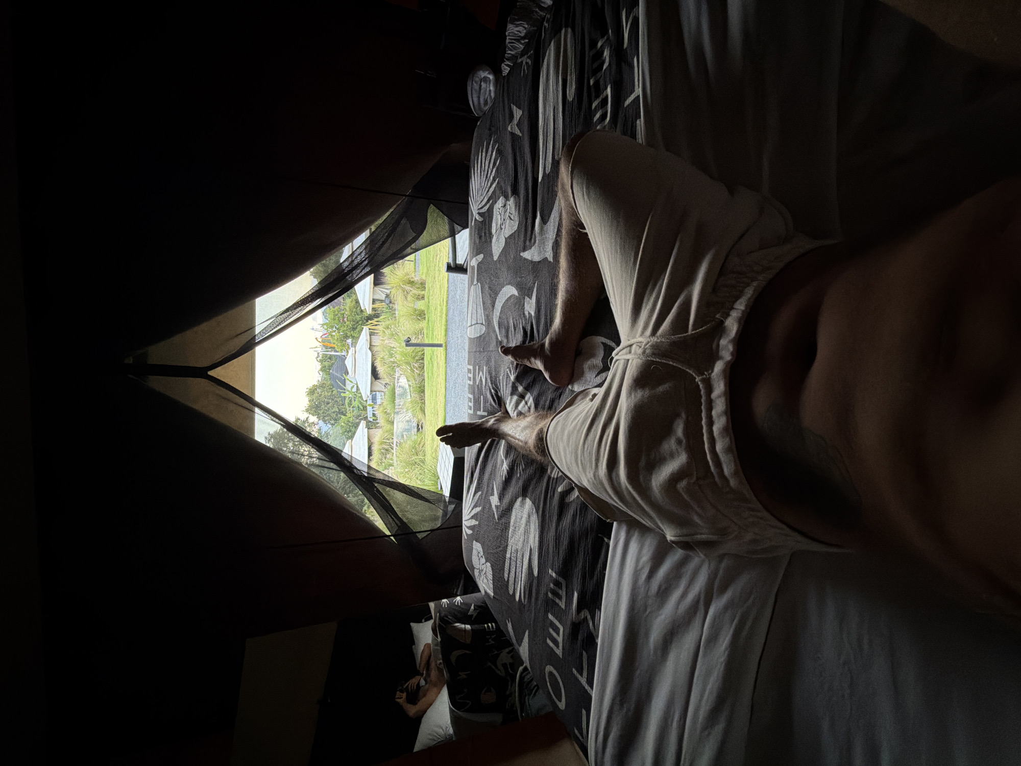 The image depicts a person lying on a bed inside a tent, with the tent flap open to reveal an outdoor scene in Kintamani, Indonesia. The person is positioned on their back, with their legs extended towards the opening of the tent. They are wearing light-colored shorts and their torso is visible. Another person is lying on a bed in the background, partially obscured by darkness. The bed in the foreground is covered with a patterned duvet. Outside the tent, there are several small buildings with conical roofs, lush green grass, and some trees. The sky is visible in the distance. The overall impression is one of a tranquil, outdoor lodging experience in a tropical setting.