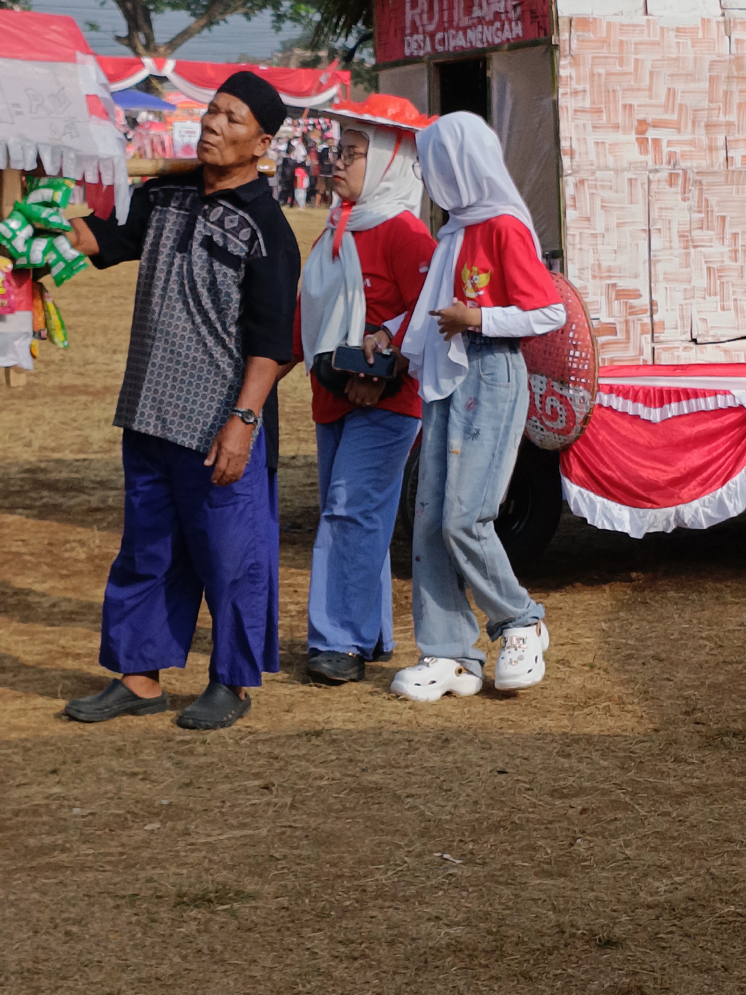 Three individuals are standing outdoors on a dry, grassy field. The person on the left is an adult male wearing a black peci hat, a black shirt with a patterned design, and loose-fitting blue pants. He is holding a bunch of green-wrapped items in his left hand. To his right stands an adult female wearing a white hijab, a red shirt, and blue jeans. She is holding a dark-colored phone in her right hand. The third person, to the far right, appears to be a young female wearing a white hijab and a red shirt with a crest on the left chest, paired with blue jeans and white clogs. Behind the individuals is a structure with a woven bamboo facade, decorated with red and white fabric draping along the bottom. A sign with text, partially obscured, is visible at the top of the structure. The ground is covered in dry grass and dirt, suggesting an outdoor event or market. The lighting indicates it is daytime.