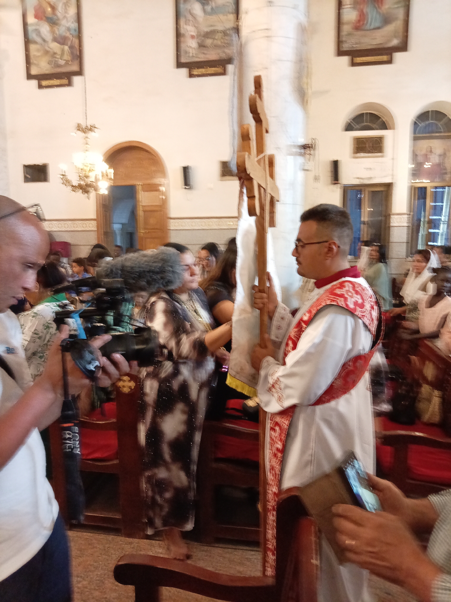 The image shows a church interior in an unknown city, with several people present. A cameraman on the left is filming a man dressed in religious garments holding a wooden cross. Another person is taking a photo with their smartphone. There are paintings on the walls, a chandelier, and rows of chairs.