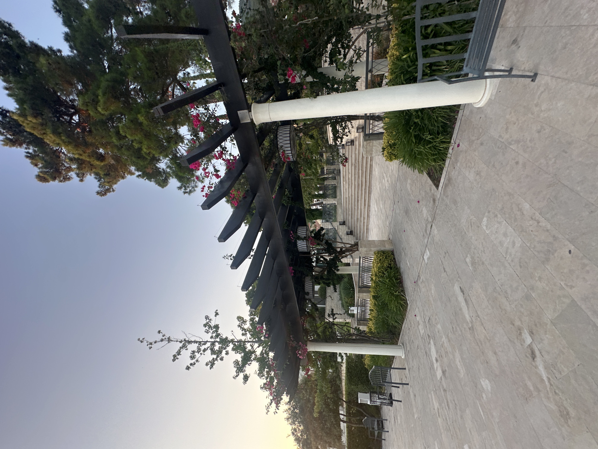 The image depicts an outdoor scene in Kumbor, Montenegro, featuring a paved courtyard with a dark wooden pergola. The pergola has white cylindrical columns supporting its structure, and it is adorned with vibrant pink bougainvillea flowers. Beyond the pergola, a wide stone staircase leads upwards, flanked by manicured green bushes and decorative iron railings. Two dark metal benches are visible in the foreground, one to the left and another to the right, positioned on the paved area. The sky is a clear, pale blue, suggesting either early morning or late afternoon light, with the sun casting a warm glow on the foliage of a large pine tree in the upper right portion of the image. The overall impression is one of a serene and well-maintained outdoor space within a resort or hotel setting.