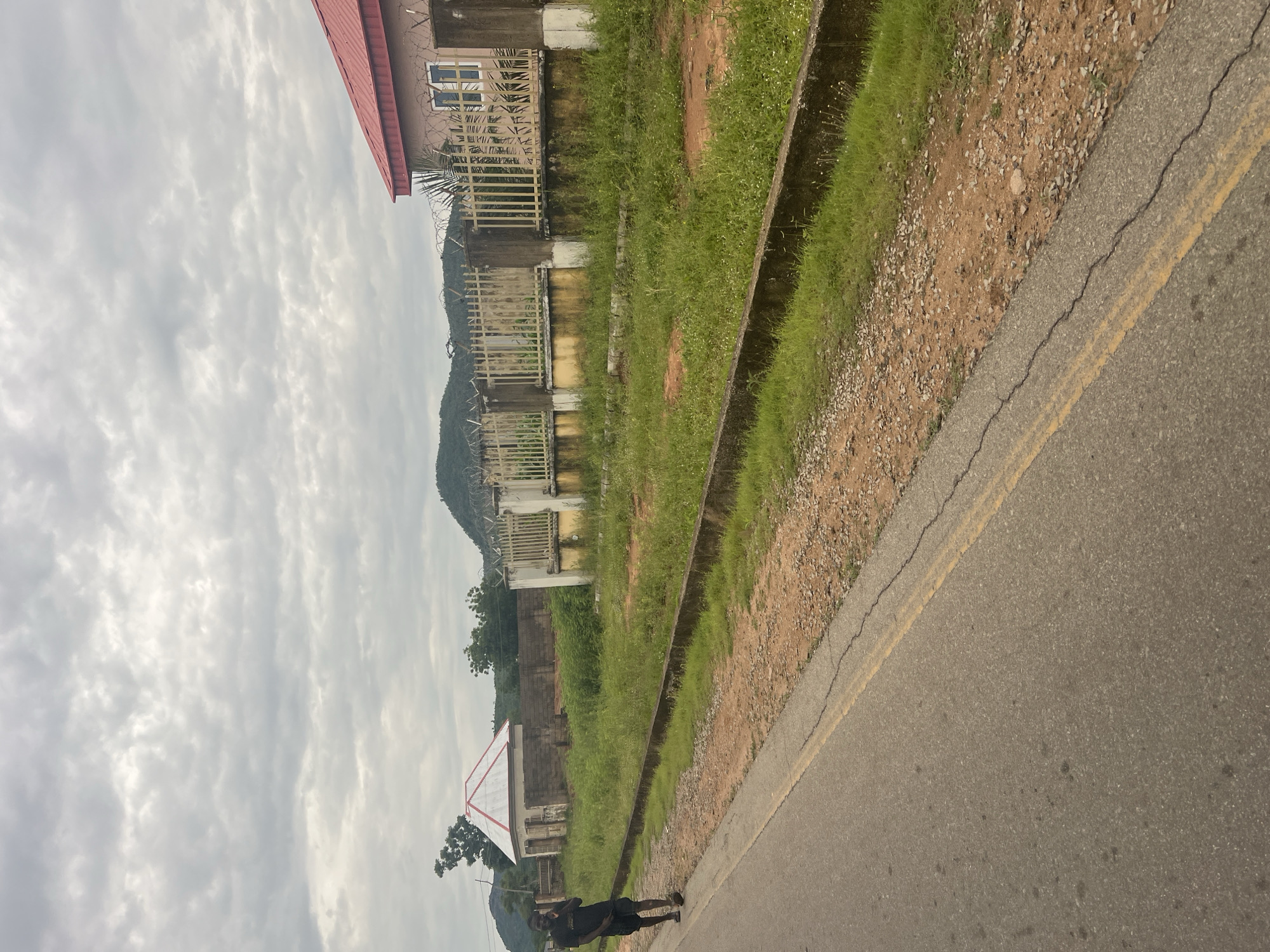 The image depicts a street scene in Kona Garu, Nigeria. A young man wearing a black t-shirt and shorts is walking on the left side of a paved road, talking on a cell phone. The road has a double yellow line in the center. On the right side of the road, there is a patch of dry, gravelly earth bordering a grassy area. Beyond the grass, there is a concrete barrier and then a chain-link fence topped with barbed wire. Behind the fence, two buildings are visible: one is a small, white structure with a red and white roof, and the other is a larger building with a red roof and light-colored walls. In the background, a green, hilly landscape is visible under a cloudy sky.