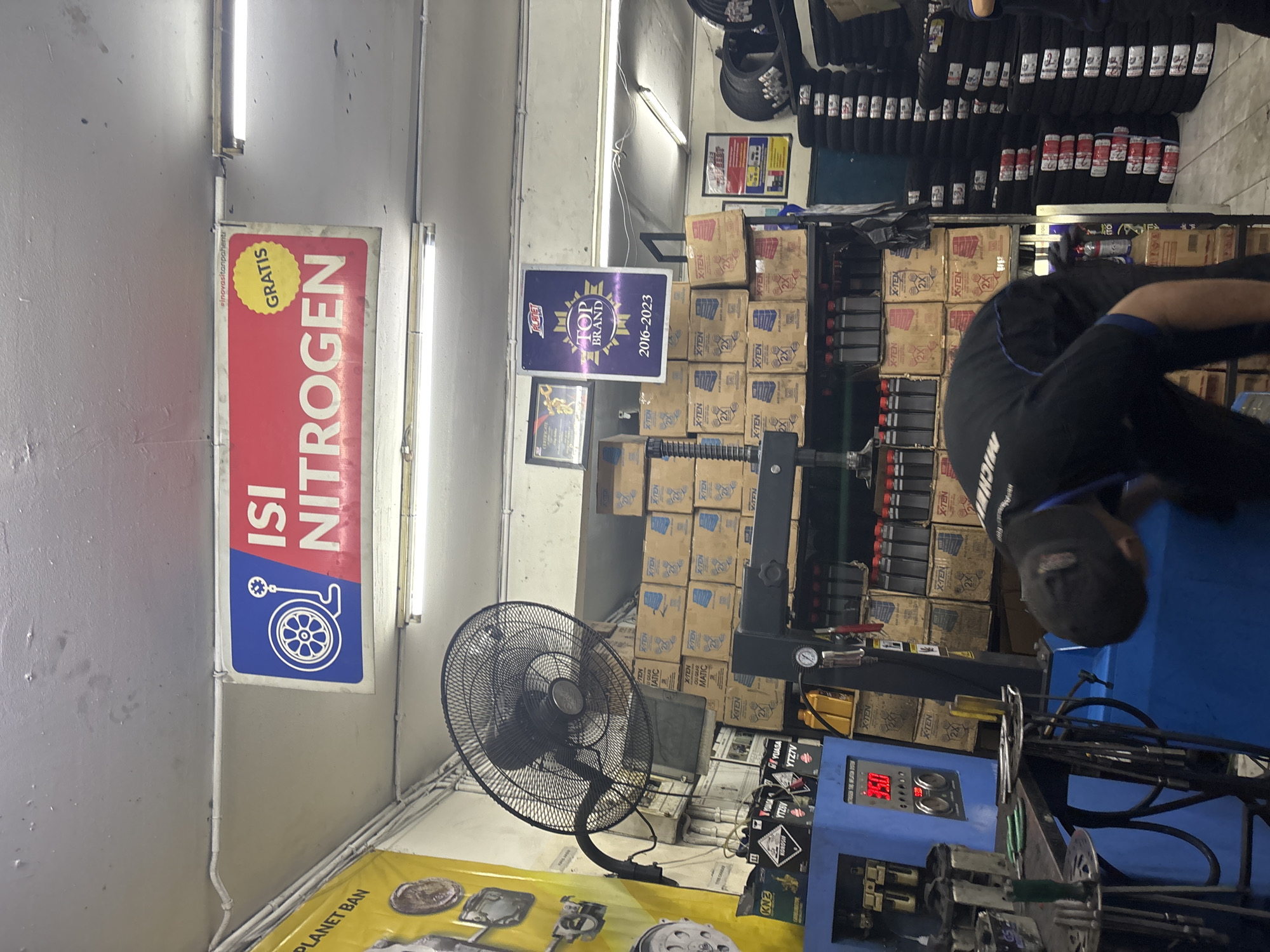 The image captures the interior of a tire shop in North Jakarta, Indonesia, focusing on a nitrogen tire inflation system. A digital display on the machine shows "95.0" and "3:00", likely indicating tire pressure and time. To the left of the machine, a yellow banner with the text "PLANET BAN" is partially visible. Above, a blue and red sign reads "ISI NITROGEN" with a graphic of a tire and a nitrogen tank, and a yellow "GRATIS" (free) sticker. Stacks of tire boxes labeled "X-TEN" are visible on shelves behind the machine, along with several bottles of oil. A large black oscillating fan is mounted on the wall. In the foreground, a person wearing a black shirt with "MICHELIN" visible on the back is bending over, appearing to work on or inspect something near the machine. Further to the right, stacks of tires are neatly arranged. The environment appears to be a well-lit workshop, suggested by the fluorescent lighting fixtures on the ceiling.