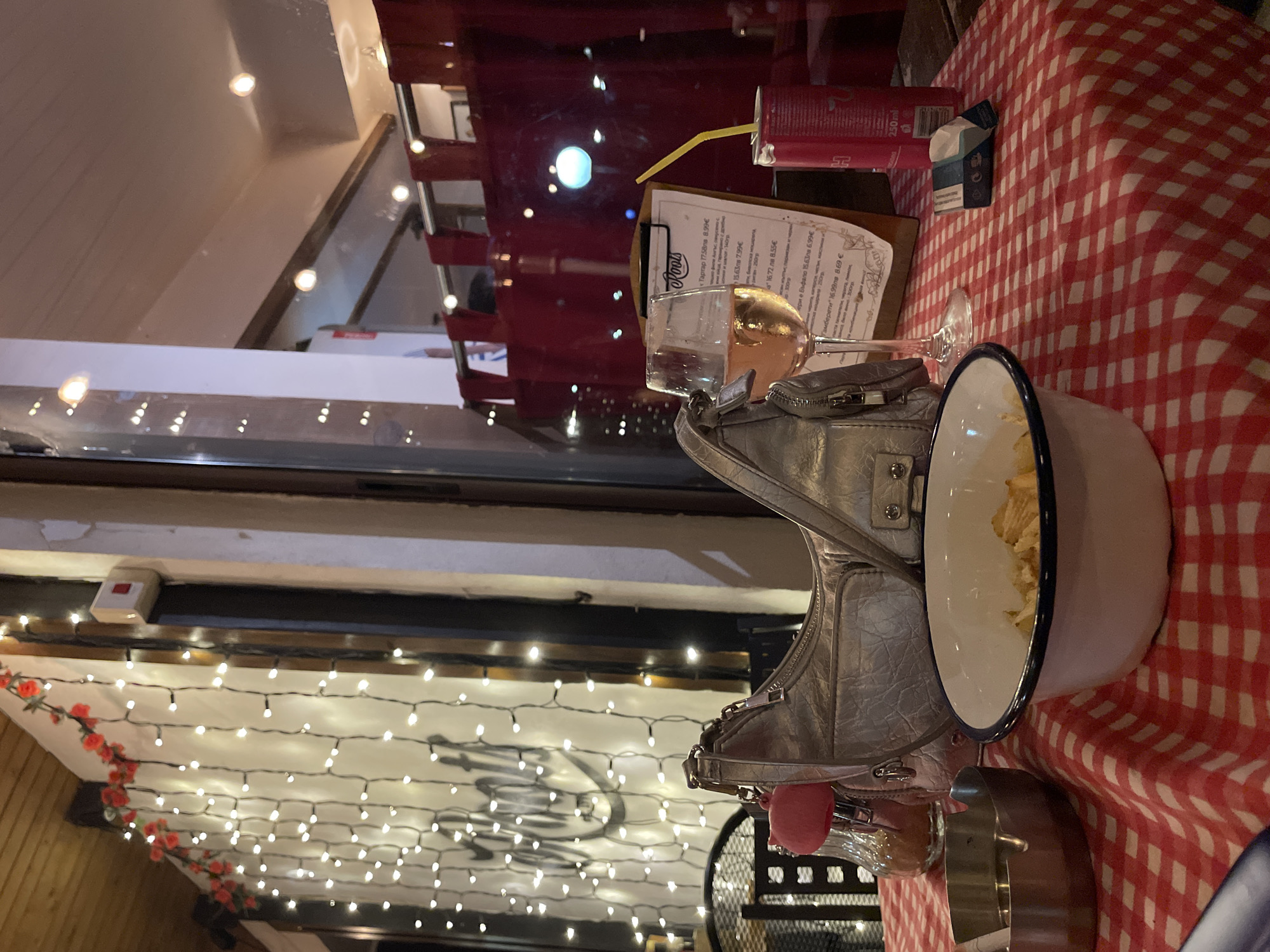 The image depicts an outdoor dining table at night in Blagoevgrad, Bulgaria. The table is covered with a red and white checkered tablecloth. Objects on the table include, from left to right: a metallic ashtray with a cigarette resting in a groove; a clear glass shaker with a pink cap containing brown granular material; a silver-grey textured handbag with a pink pig-shaped charm attached; a white enamel bowl with a blue rim holding light-colored snacks (likely potato chips); a clear wine glass containing pale rosé wine; a wooden clipboard menu with "Roots" prominently displayed at the top and additional text in Bulgarian, listing items and prices; a red 250ml beverage can with a yellow straw inserted; and a partially open blue and white cigarette packet. The background features vertically strung white fairy lights intertwined with red artificial roses, illuminated against a light-colored wall or partition. Reflections of an interior space, including red drapery and overhead lights, are visible in a window behind the table setup. No individuals are present in the frame.