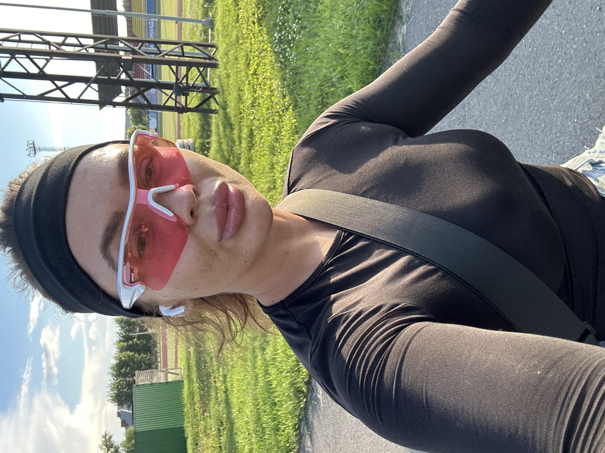 The image depicts a woman wearing a black long-sleeved shirt, a black headband, white wireless earbuds, and pink-tinted sports sunglasses. She appears to be outdoors, with a grassy field and a paved path visible. In the background, there are some trees, a green metal fence, and a tall metal structure that resembles stadium lighting or a sports facility. The sky is blue with some clouds. The woman is looking towards the camera. The scene is consistent with a location in Yuzhnouralsk, Russia, possibly near a sports complex or stadium.