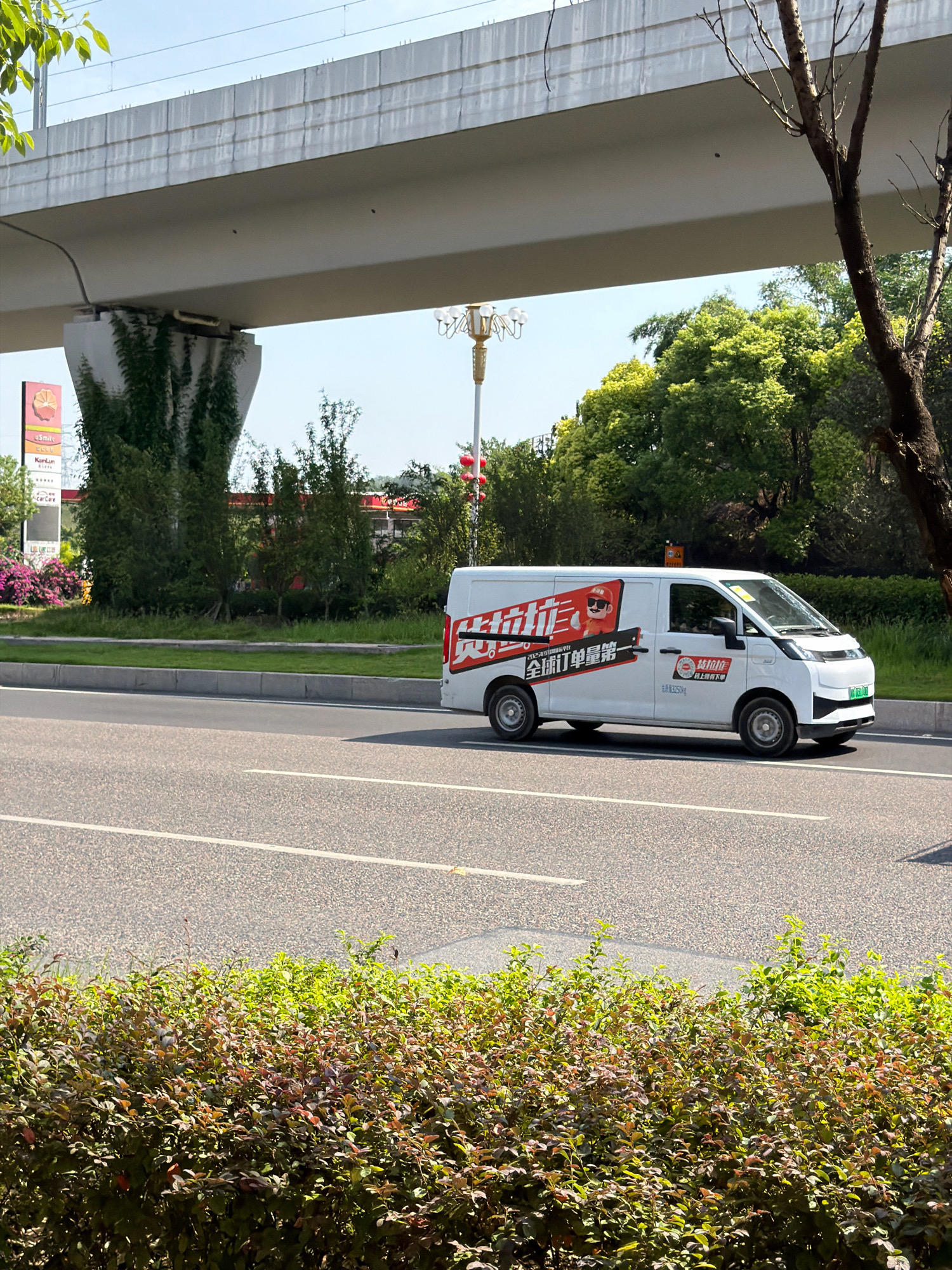 A white electric delivery van is driving on a road in Jiangjin District, Chongqing, China. The van has red and white graphics on its side, including a cartoon character wearing sunglasses and the text "全球订单量第一" (Global order volume number one). A portion of an elevated structure, likely a bridge or overpass, is visible in the upper part of the image, with a concrete pillar supporting it. Lush green trees and some flowering bushes are present in the background and foreground, indicating an outdoor, possibly suburban or park-like environment within the city. The sky is clear and blue. The van is the primary subject, captured in motion on a paved road with visible lane markings.