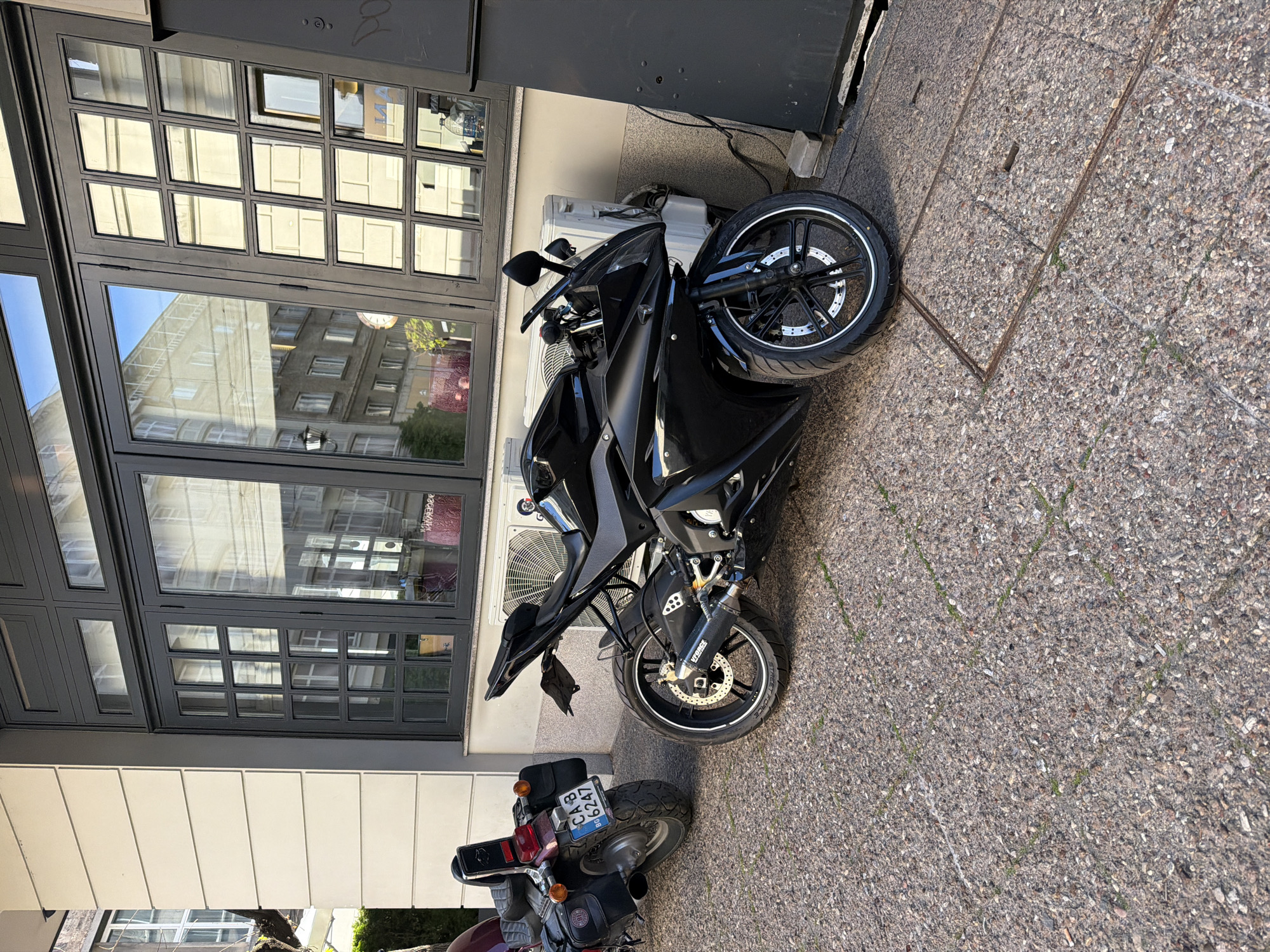 An outdoor scene captures two motorcycles parked alongside a building on a sunny day. The primary subject is a sleek, black sports motorcycle with dark wheels and disc brakes, positioned on a light gray, coarse paved surface. Behind the front wheel of the black motorcycle, a white outdoor air conditioning unit is visible, mounted against a building.
The building features large, dark-framed windows that reflect the bright blue sky and the facade of another building across the way. Below the windows, the building has a cream-colored wall with vertical paneling.
Partially visible in the lower left of the frame, behind the black motorcycle, is the rear section of an older, red or maroon motorcycle. This motorcycle's license plate, "CA 6247 BH," is clearly legible, indicating a registration from Sofia, Bulgaria. Text like "VERSYS" is partially visible on the black motorcycle's suspension or exhaust, and "STREET" or "STREE" can be seen on its tire sidewall. The paved ground displays a subtle grid pattern. The clear reflections and distinct shadows suggest it is daytime with bright sunlight. No individuals are present in the image.