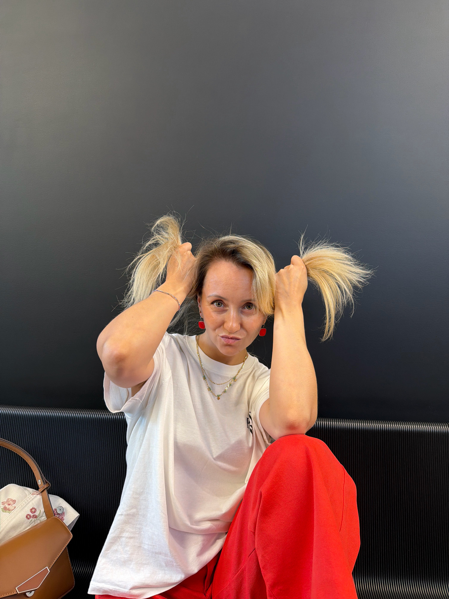 A young blonde woman with bright red square earrings and a white t-shirt is posing for a photo, holding her blonde pigtails. She is wearing red pants and sitting on a black bench in front of a black wall. A brown handbag is visible on the left side of the frame. The image was taken indoors, and there are no recognizable landmarks or signs to indicate a specific location in Izhevsk, Russia.