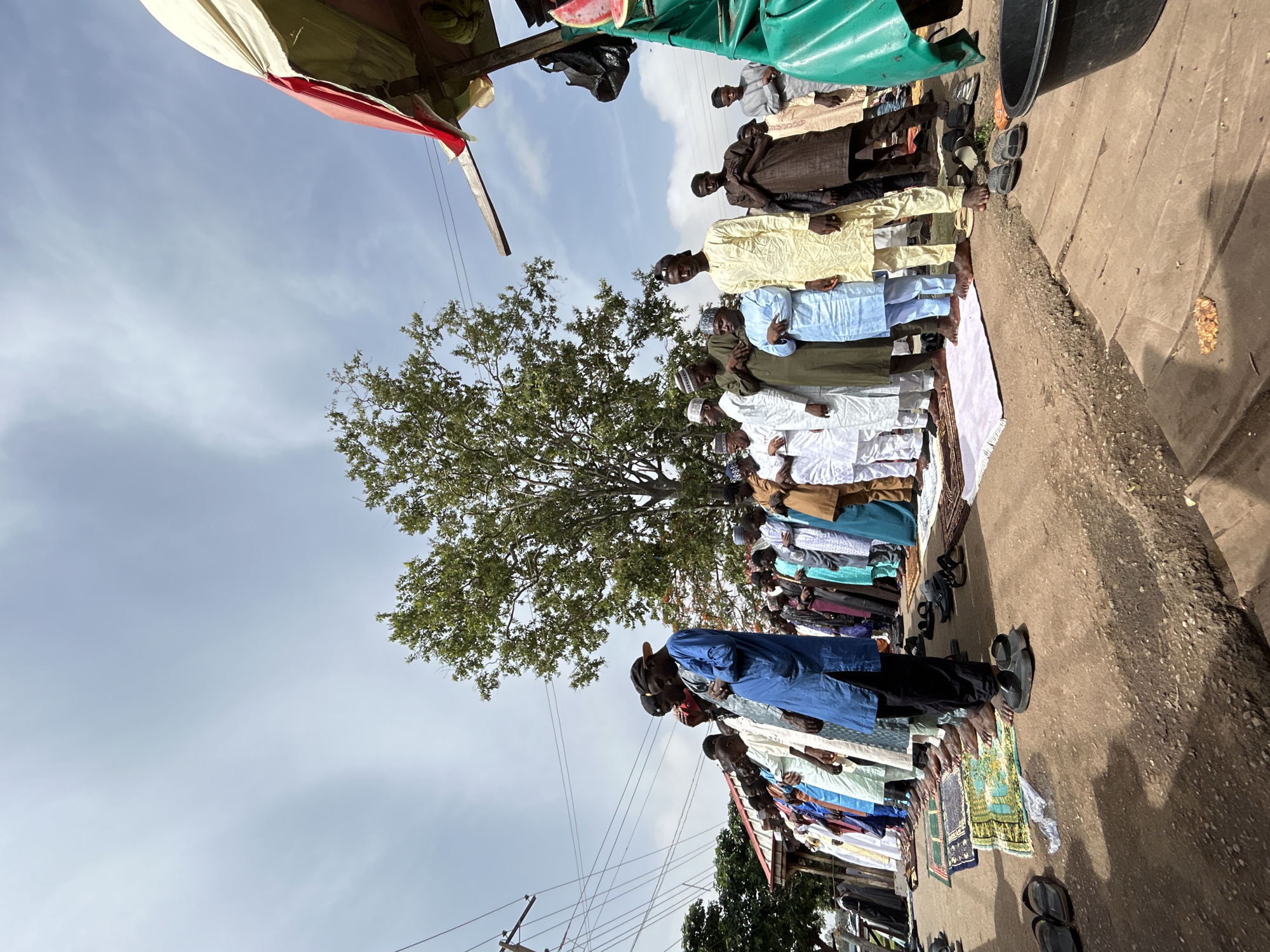 In Jos, Nigeria, a group of men is praying outside. The men are lined up, facing the same direction, on a concrete surface. Most of the men are wearing traditional clothing, varying in color from white to blue to yellow to brown. Some men have removed their shoes, which are placed nearby. A tree is visible in the background. The sky is overcast. Power lines run overhead.