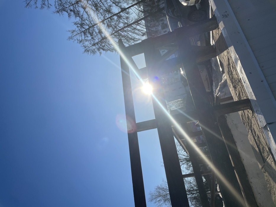 The image captures an outdoor scene in Chaikovsky, Russia, under a clear, bright blue sky with direct sunlight. The sun, positioned in the upper right quadrant, emits a strong glare and prominent lens flares. A dark-colored wooden structure, resembling a railing or fence, spans the lower and mid-section of the frame, partially silhouetted against the sky and sunlight. Below this structure, a light-colored, possibly white, horizontal surface is visible, covered with dirt and patches of snow. In the background on the left, several bare deciduous trees are present. On the right, a cluster of tall, slender conifer trees stands, beyond which a multi-story building and several parked vehicles are discernible on what appears to be a street or parking area. Patches of snow and disturbed earth are visible on the ground, indicating a winter or early spring setting. No individuals are depicted.