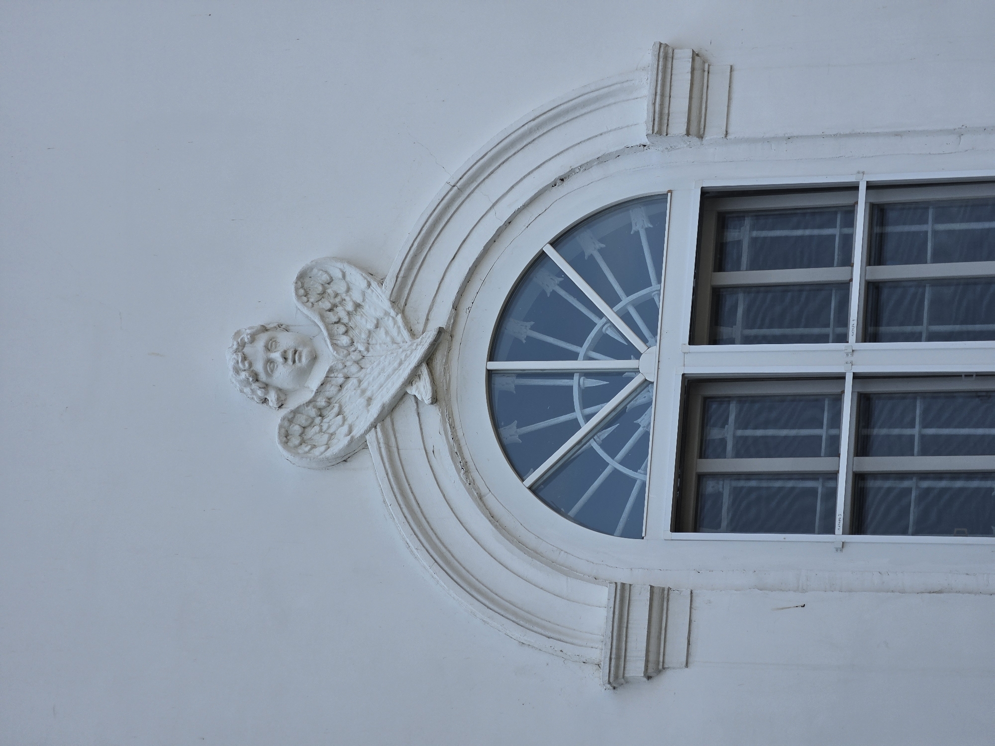 The image shows a close-up, eye-level view of a white stucco building facade in Nizhny Novgorod, Russia. The focus is on an arched window topped with a decorative plaster element featuring a cherub's head with outstretched wings. The window itself is divided into a semi-circular fanlight with a white radial grille, above a rectangular section divided into two panes horizontally and three panes vertically by white mullions. The panes are reflective, showing a blue sky. The plaster decoration above the window has some minor cracks and weathering. Below the window, there are horizontal bars and some faint writing that appears to be in Cyrillic. The background is a plain, slightly textured white wall.