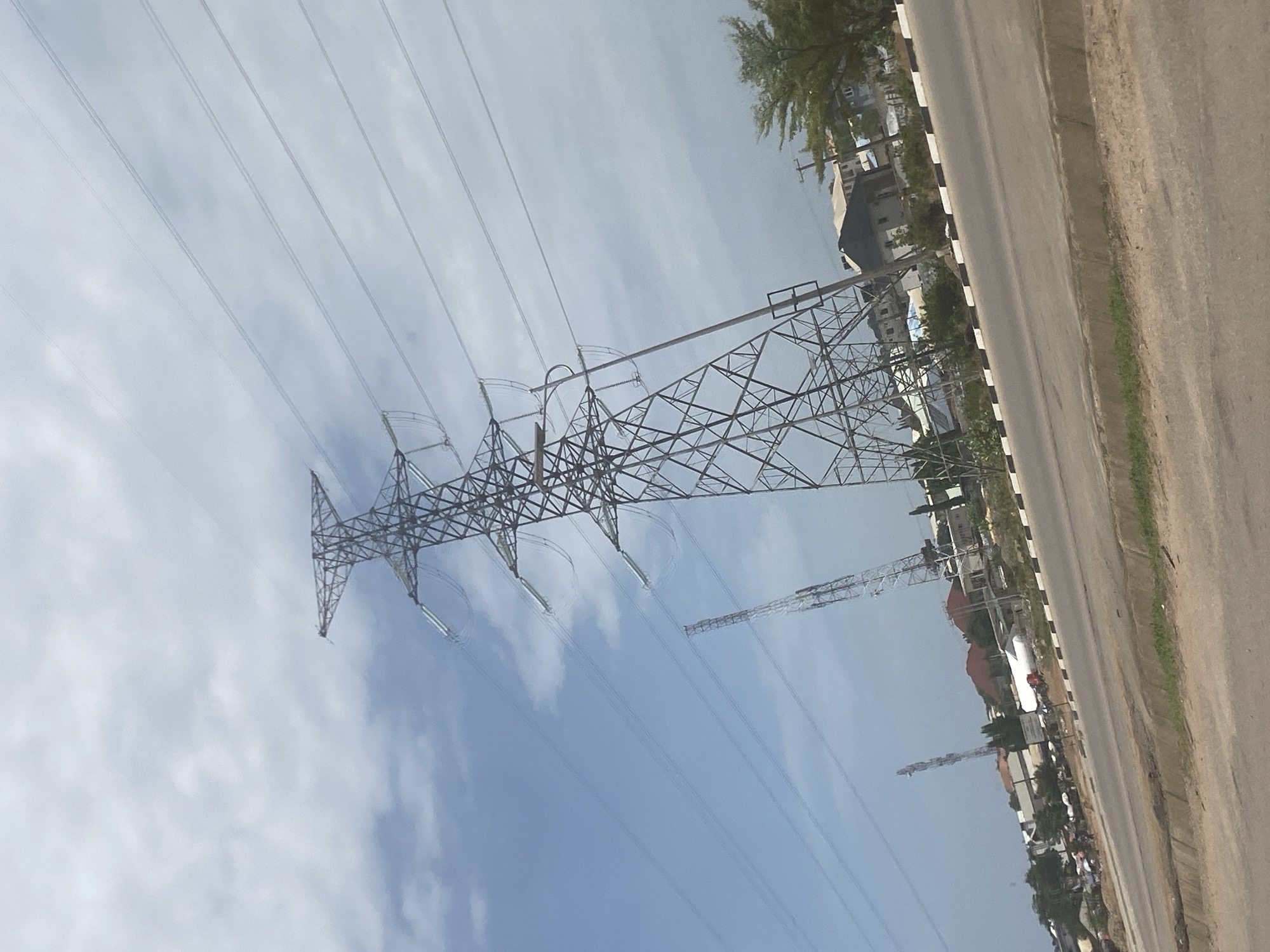 The image captures an overhead view of an electricity transmission tower in Abuja, Nigeria. The metal tower is seen against a backdrop of a cloudy, blue sky, with multiple power lines extending from the tower. Alongside the tower, a road runs parallel, with a painted median strip. Buildings and trees are visible on the other side of the road. Further in the background, another tower and antenna are visible.