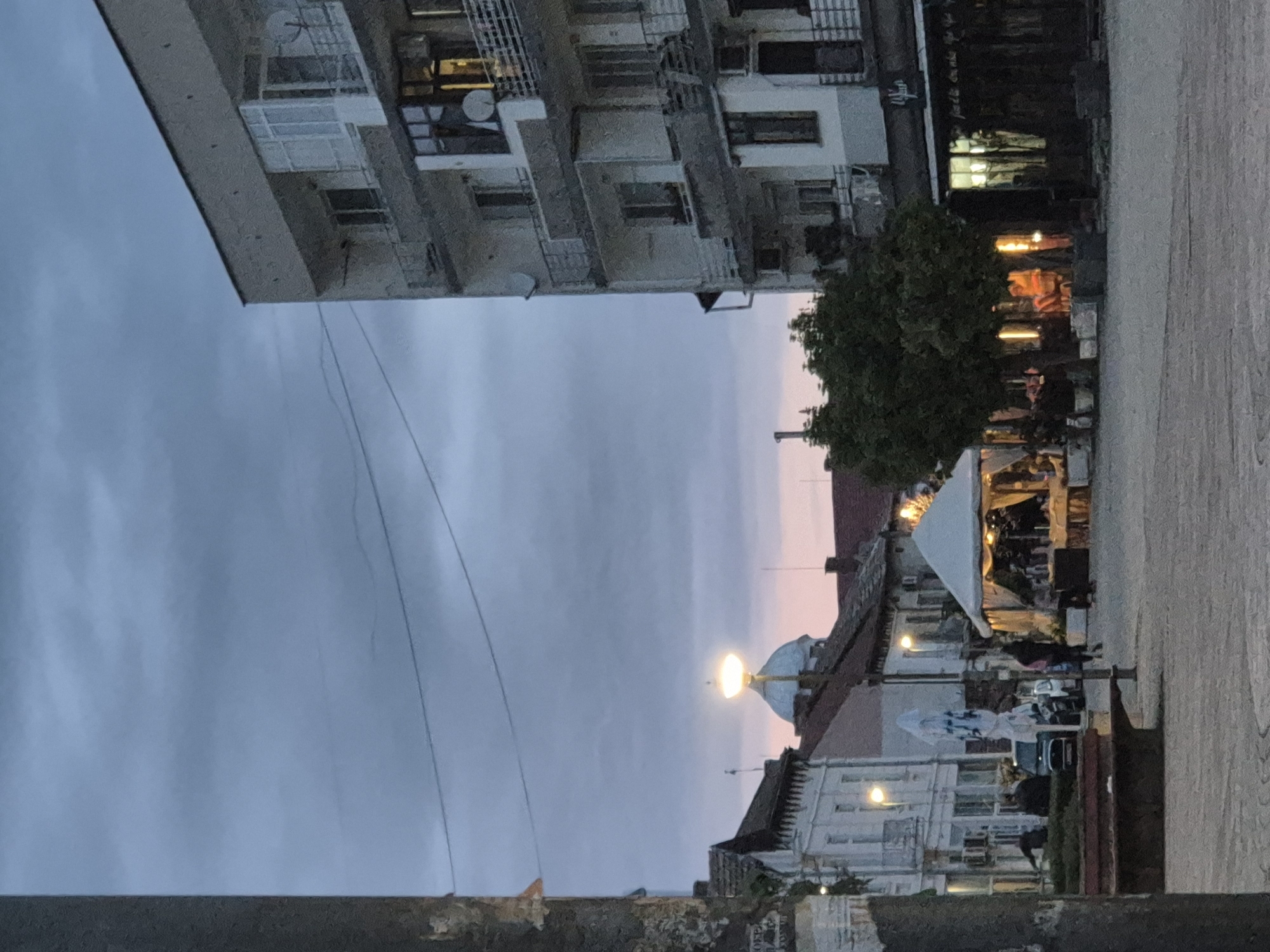 The image shows a street scene in Pavlikeni, Bulgaria. A tall apartment building is on the left, with several floors visible. On the right side, there are smaller buildings, including a building with a dome illuminated by a light. There is a tree in the middle. A street runs between the buildings, and people are present in the scene. The sky is cloudy, with some pink hues at the horizon. A barrier or ledge is visible in the foreground.
