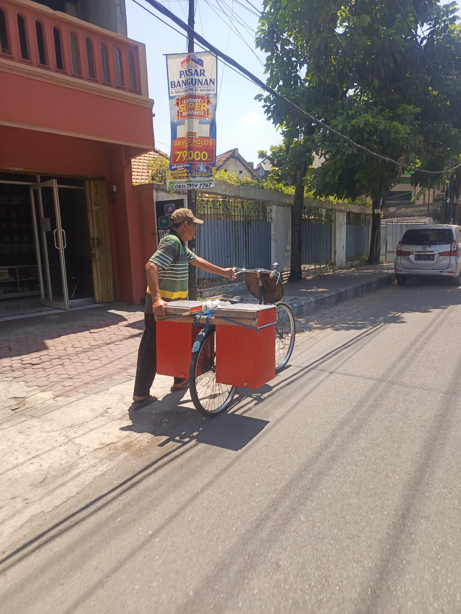 A man in a striped shirt and cap stands next to a red cargo bicycle on a street in Bebekan, Indonesia. The bicycle has two large red storage boxes and a brown basket. The man is holding onto the handlebars, and appears to be engaged in activity related to the bicycle or its cargo. In the background, there is a building with an orange facade, a banner advertising building materials, trees, a fence, and a silver van. The scene is illuminated by bright sunlight, casting distinct shadows.