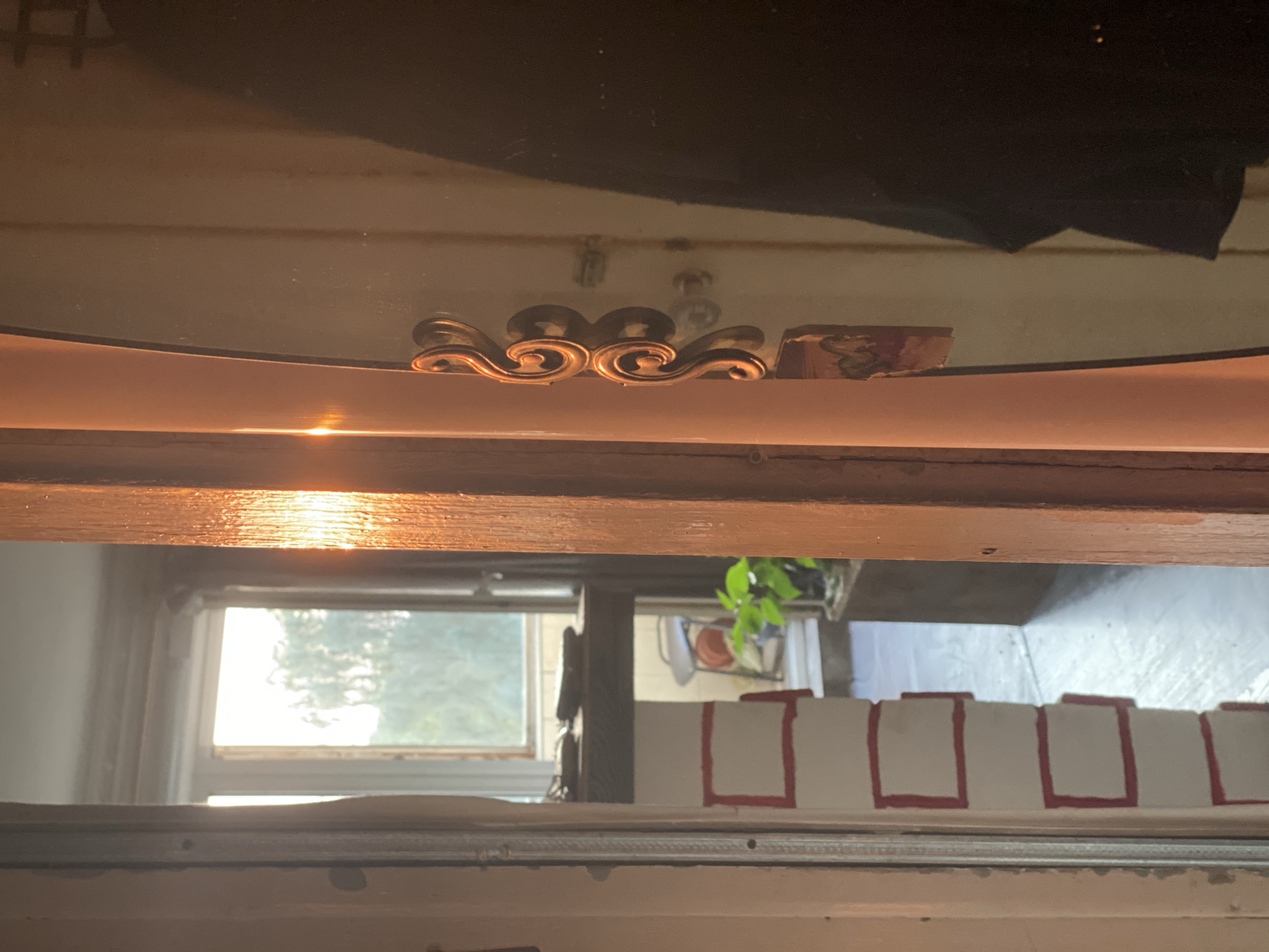 The image depicts an interior view looking towards a window through an open doorway. The sunlight entering the frame creates a strong glare on a wooden doorframe and a reflective surface. In the foreground, a wooden doorframe is visible on the right, with a narrow, light-colored pillar on the left. Through the doorway, a window with a view of trees can be seen. Below the window, a dark shelf holds a few objects. To the left of the shelf, a white column is decorated with red rectangles. A potted plant is visible in the background. On the reflective surface to the right, there is a decorative golden handle. The overall impression is of an indoor space with natural light. The location is identified as Novokuznetsk, Russia.