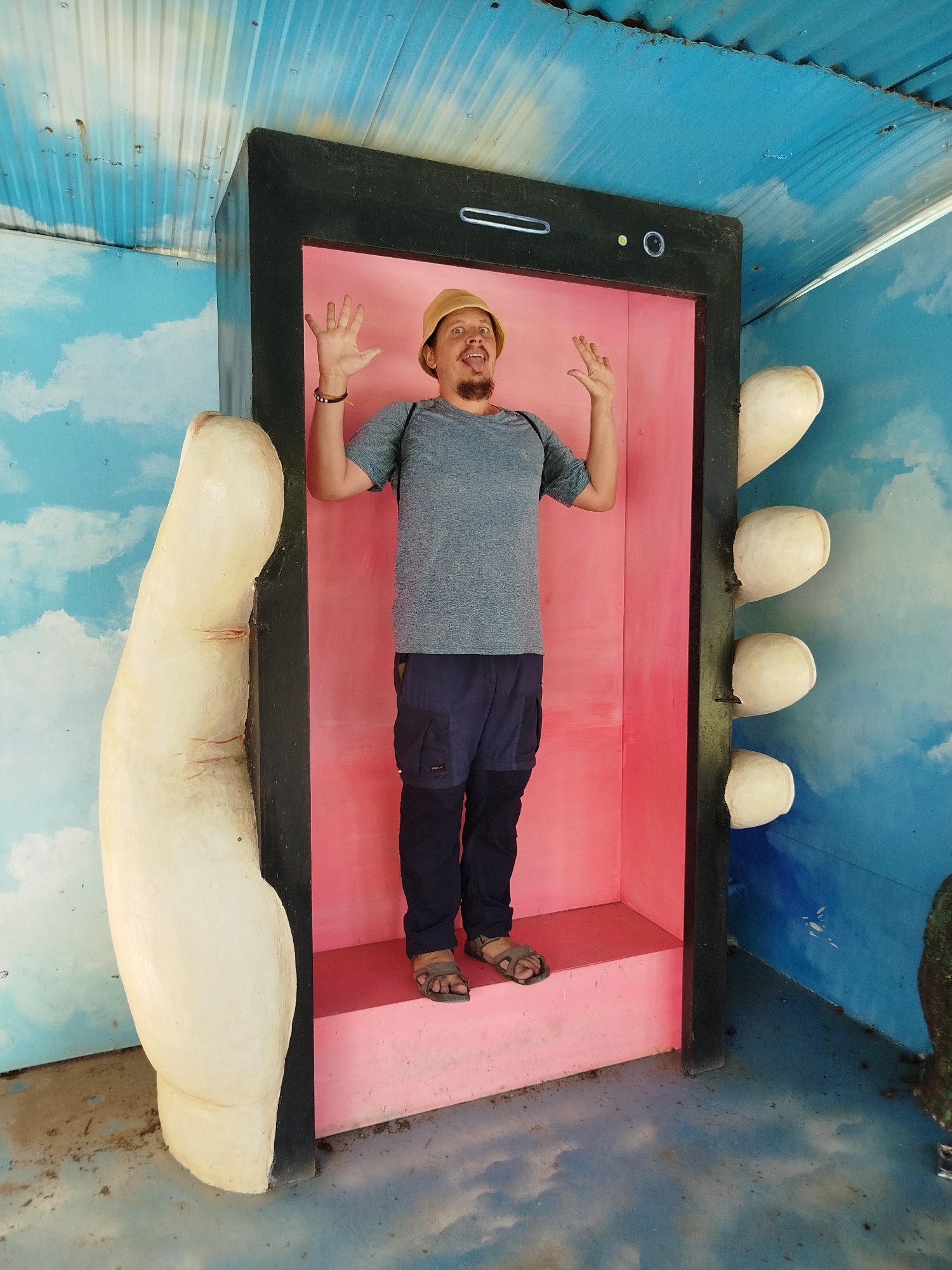 A medium shot shows a man posing within a large, fabricated smartphone-like structure outdoors. The man wears a grey t-shirt, dark blue cargo pants, brown sandals, and a tan bucket hat. He stands with both hands raised, fingers spread. The smartphone prop is black with a pink interior, which functions as a standing platform for the man. The structure has painted features mimicking a phone's front camera and speaker. To the side of the "phone" is a large, sculpted hand. The backdrop and surrounding environment depict a painted sky and clouds, with hints of greenery at the edges of the frame. The picture was taken in Da Lat, Vietnam.