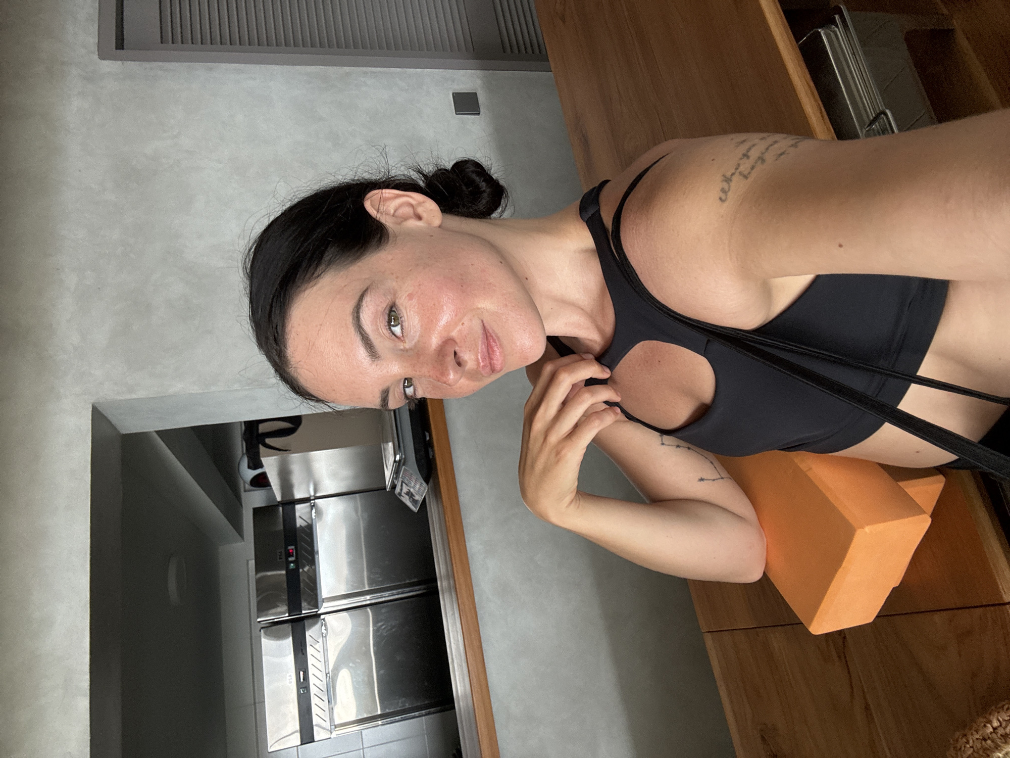 In North Kuta, Indonesia, a woman with dark hair pulled into a bun takes a selfie indoors. She is wearing a black sports bra and leans on her left arm, which rests on an orange yoga block. Her right hand is near her neck. Behind her, a wooden counter separates her from a kitchen area with stainless steel appliances and a gray wall. A door with horizontal slats is visible to her right. She has a tattoo on her right arm and freckles on her face.