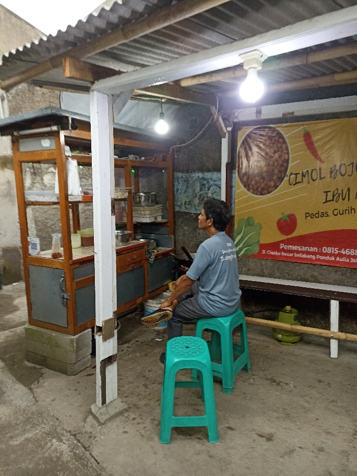 The image shows a man sitting on a green plastic stool in front of a food stall in Jatinangor, Indonesia. The stall is made of wood and has a glass display case. The man is wearing a blue shirt and gray pants. He is looking to his right. The stall has a sign that says "Cimol Bojo Ibu" and has a phone number for orders. There are two green plastic stools in the image, one of which is occupied. The stall is located in a small, open-air area with a concrete floor. The stall is located in front of a wall with a sign on it. There is a wooden beam above the stall. The image was taken in Jatinangor, Indonesia (latitude: -6.934, longitude: 107.775).