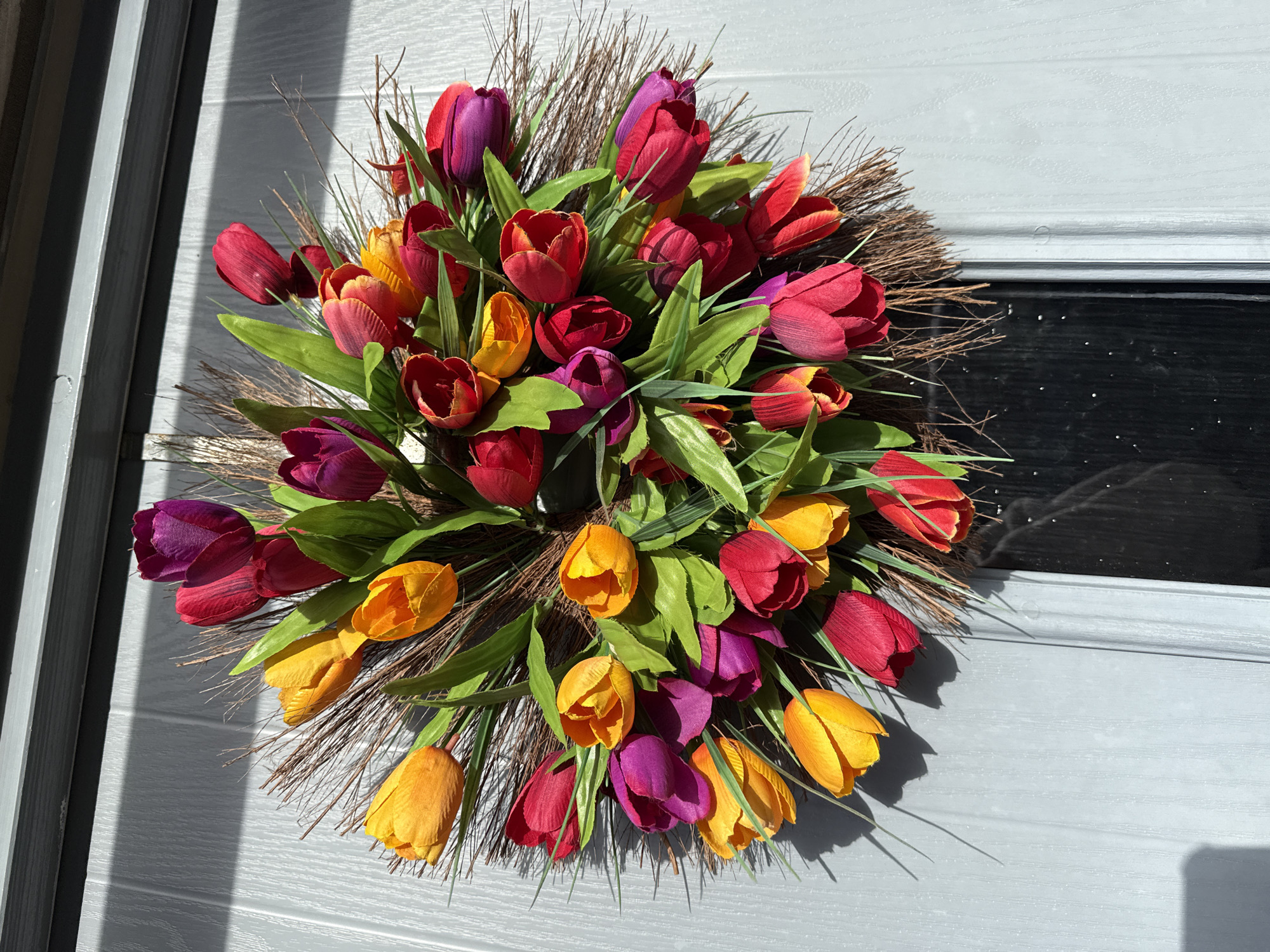 A floral wreath of tulips in shades of red, orange, and purple, with green leaves and sprigs of brown sticks, is centered on a gray door with vertical paneling on the left side and a rectangular inset on the right. The scene is bathed in sunlight, casting shadows on the door and wreath. There is no information about the city of Great Yarmouth, United Kingdom in the image.