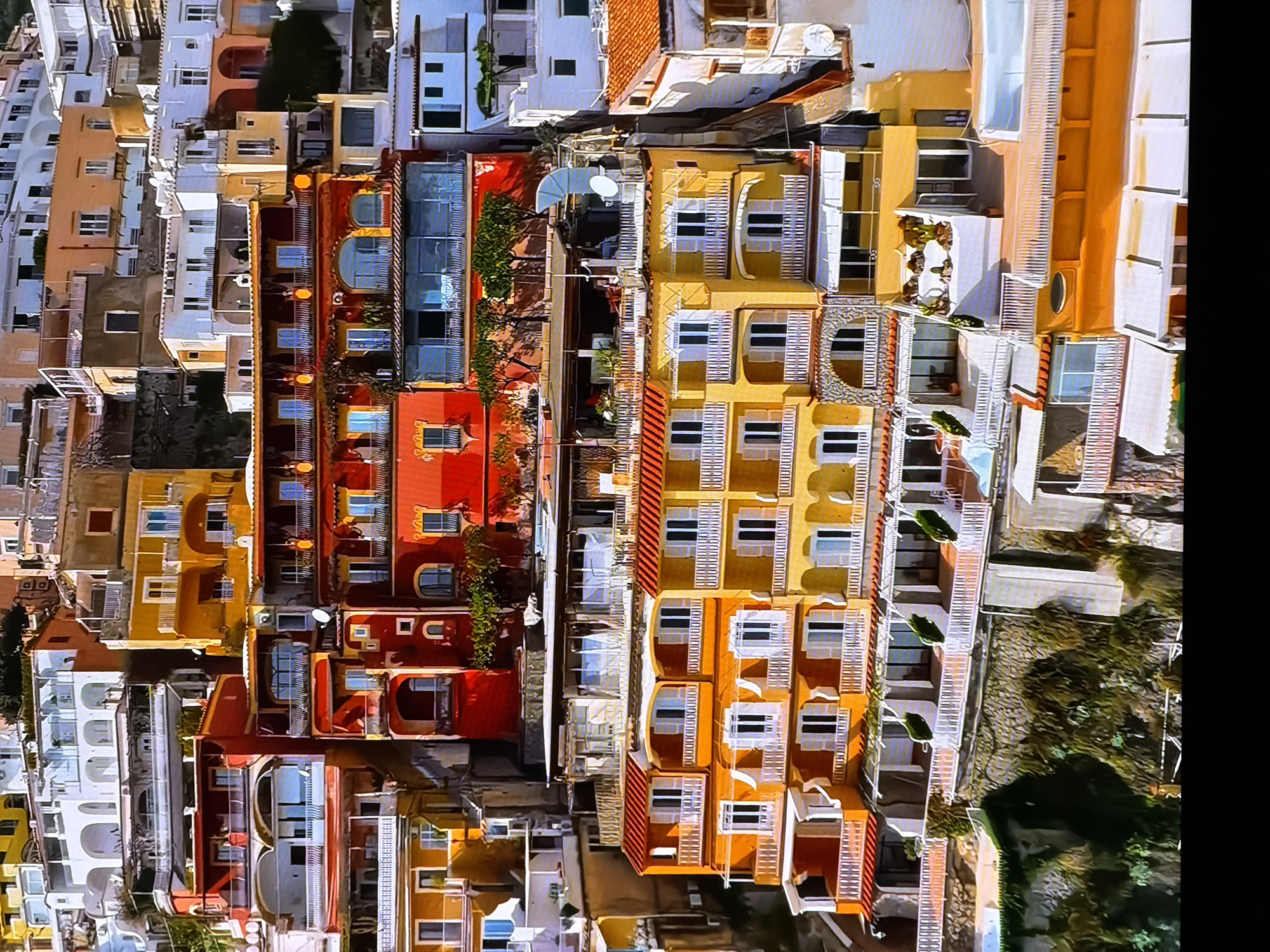 The image depicts a colorful, multi-story cliffside village with buildings of varying architectural styles and vibrant colors, predominantly orange, yellow, and red. The buildings are densely packed and built into the hillside, with many balconies, terraces, and awnings. Lush green vegetation is visible cascading down the slopes and on some of the terraces. The scene is bathed in sunlight, suggesting a warm climate. The specific location mentioned, Chirchik, Uzbekistan, is inconsistent with the visual characteristics of the image, which strongly resembles a Mediterranean coastal town, such as Positano, Italy. There are no discernible people or specific actions occurring in the image. The environment is characterized by steep terrain and numerous structures integrated into the landscape.