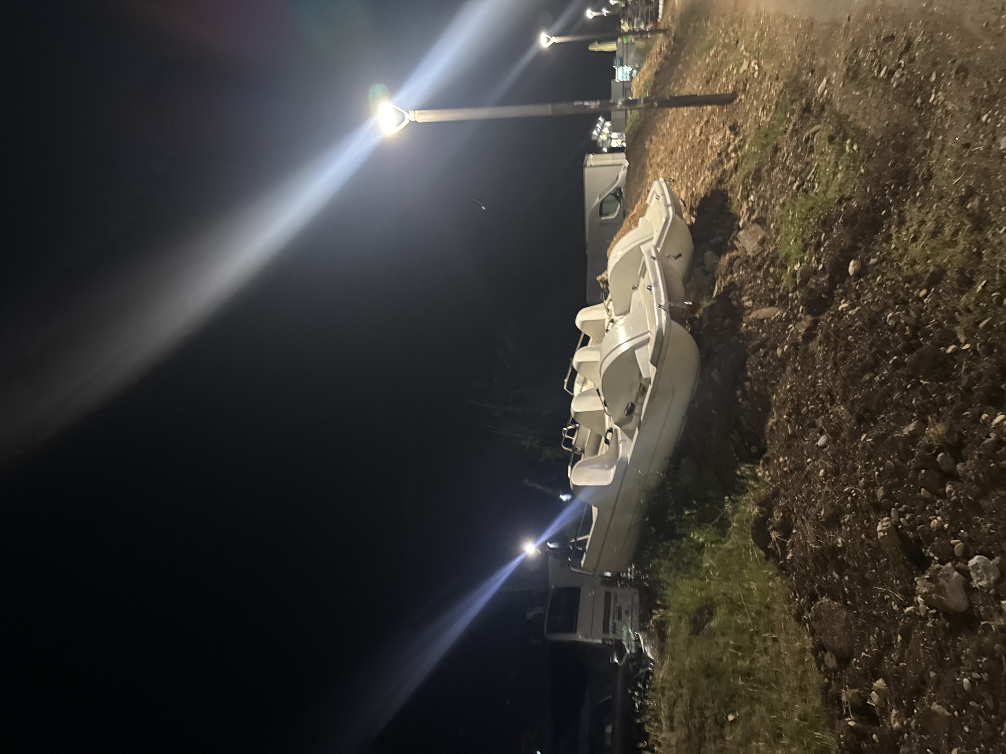 At night, in Budva, Montenegro, on a grassy incline next to a paved area, several white pedal boats are parked. One of the boats is particularly prominent, situated on the left side of the frame, with its front end angled slightly upwards. To the left of the pedal boats, a white delivery truck is partially visible, along with what appears to be a dark car. In the background, there are more pedal boats and some buildings. The scene is illuminated by streetlights, with strong beams of light cutting across the dark sky. The ground is covered with dry leaves and loose soil.