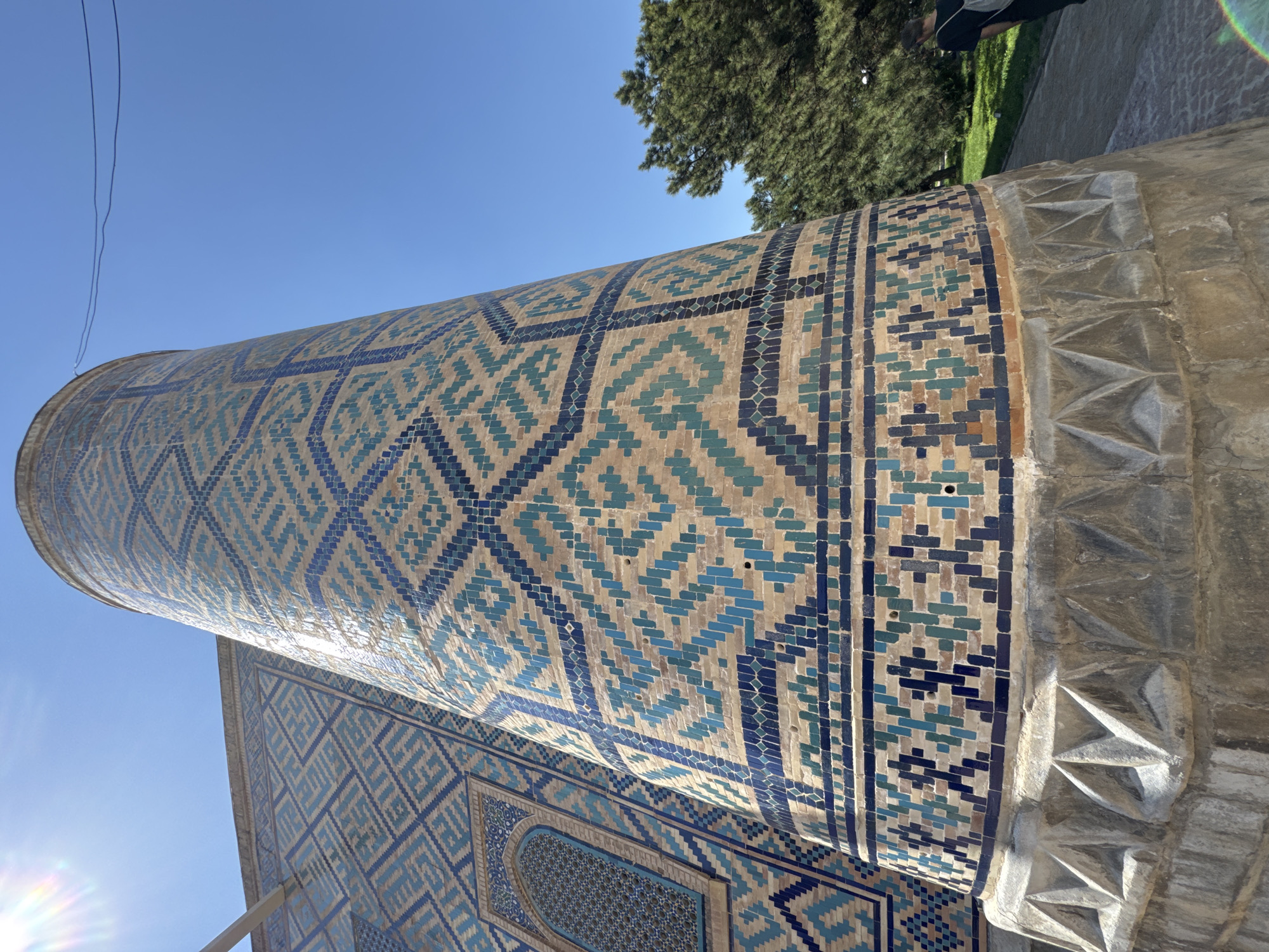 The image depicts a portion of the exterior of the Gur-e-Amir mausoleum in Samarkand, Uzbekistan. The focus is on a cylindrical column or minaret adorned with intricate mosaic tilework in shades of blue, turquoise, and tan. The tilework forms geometric patterns. Adjacent to the column is a wall also decorated with similar mosaic designs. A glimpse of a tree's foliage and a partial view of a person's leg are visible in the upper right corner of the image. The sky is clear and blue.