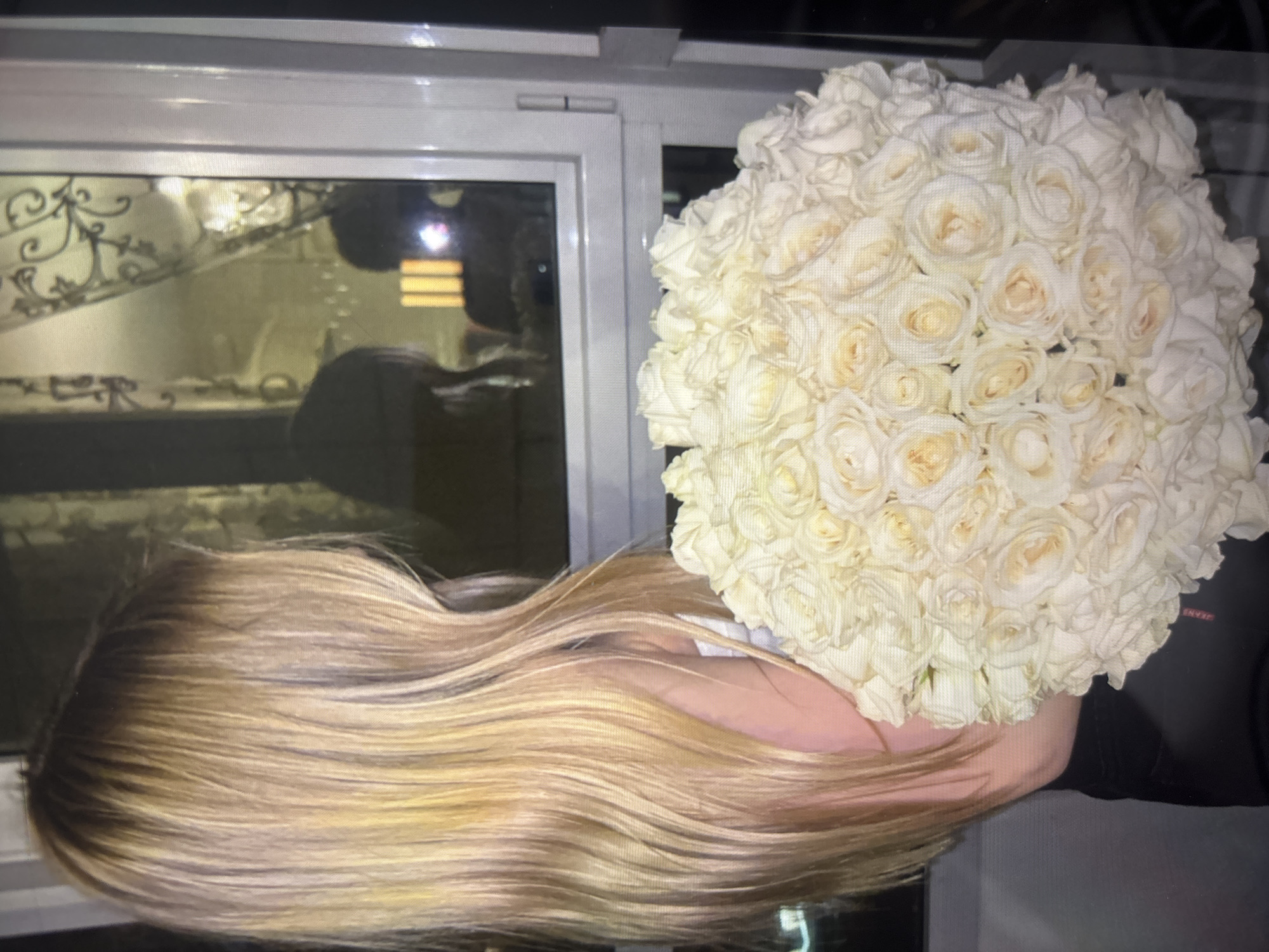 The image shows a woman with long, wavy blonde hair holding a bouquet of white roses. The woman's hair appears to have dark roots that blend into a lighter blonde color, and her hand is visible holding the bouquet. The bouquet is large and dense, composed entirely of tightly packed white roses.
Behind the woman and the roses, there's a window reflecting an interior space. The reflection reveals a staircase with ornate metal railings. The location of the photo is in the city of Unknown, Unknown.