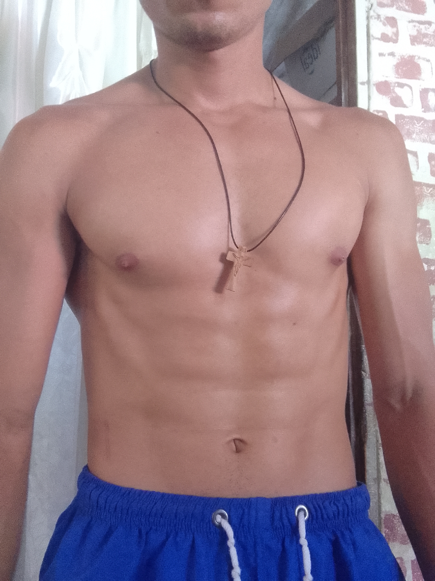 A shirtless man wearing blue shorts and a brown leather necklace with a wooden cross pendant stands in a room. The background features a white curtain and a brick wall. The location is not discernable from the image.