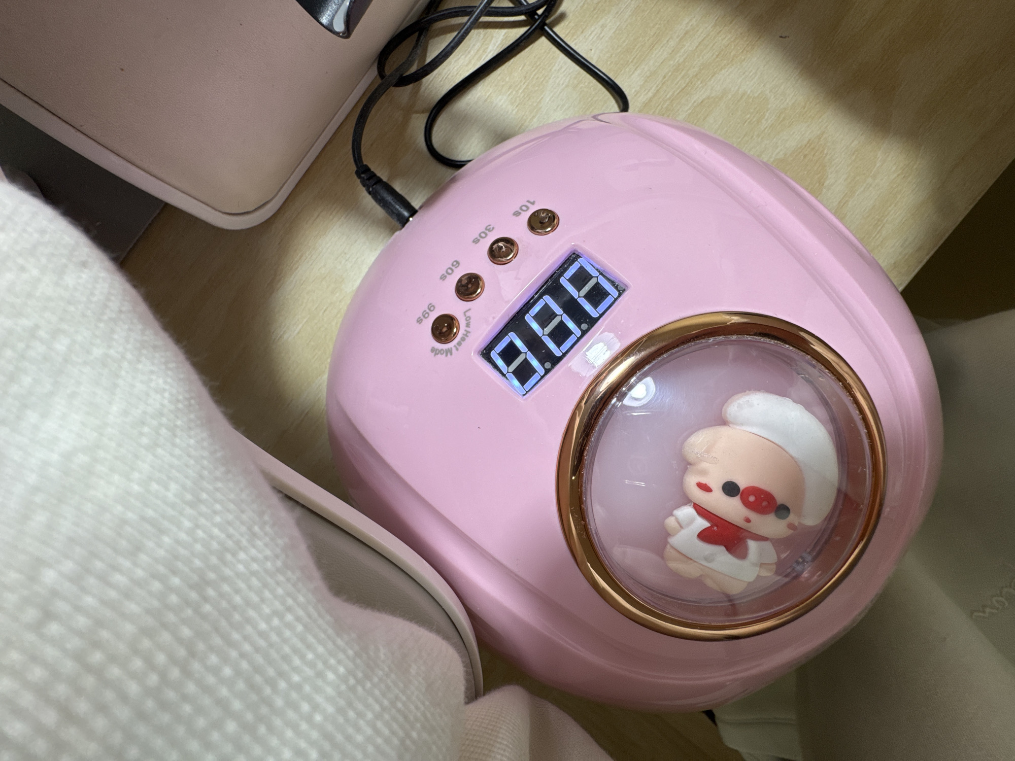 A high-angle, close-up shot displays a pink nail lamp on a light-colored wooden surface, likely a desk or floor, in an indoor setting in Bar, Montenegro. The central subject is a glossy pink nail dryer, equipped with several golden buttons marked "10s," "30s," "60s," and "99s Low Heat Mode," indicating different drying time options and a low heat mode. A bright blue digital LED display shows "8.8.8," suggesting the device is powered on. A black power cable extends from the back of the lamp onto the wooden surface.
A distinctive feature of the lamp is a small, clear, spherical enclosure on its right side, housing a cute, cartoon-style pig figurine. The pig, with its light pink complexion and prominent snout, is dressed as a chef, complete with a white hat, white jacket, and a red scarf.
To the left of the nail lamp, a soft, white, textured fabric, possibly a sweater or blanket, occupies the foreground. Part of a larger, beige electronic device or piece of furniture is visible in the upper left background. The lighting in the image is even, suggesting an indoor environment without strong shadows. Faint, indecipherable text is barely visible on a surface to the right of the lamp. The scene evokes a personal care or beauty routine setup.