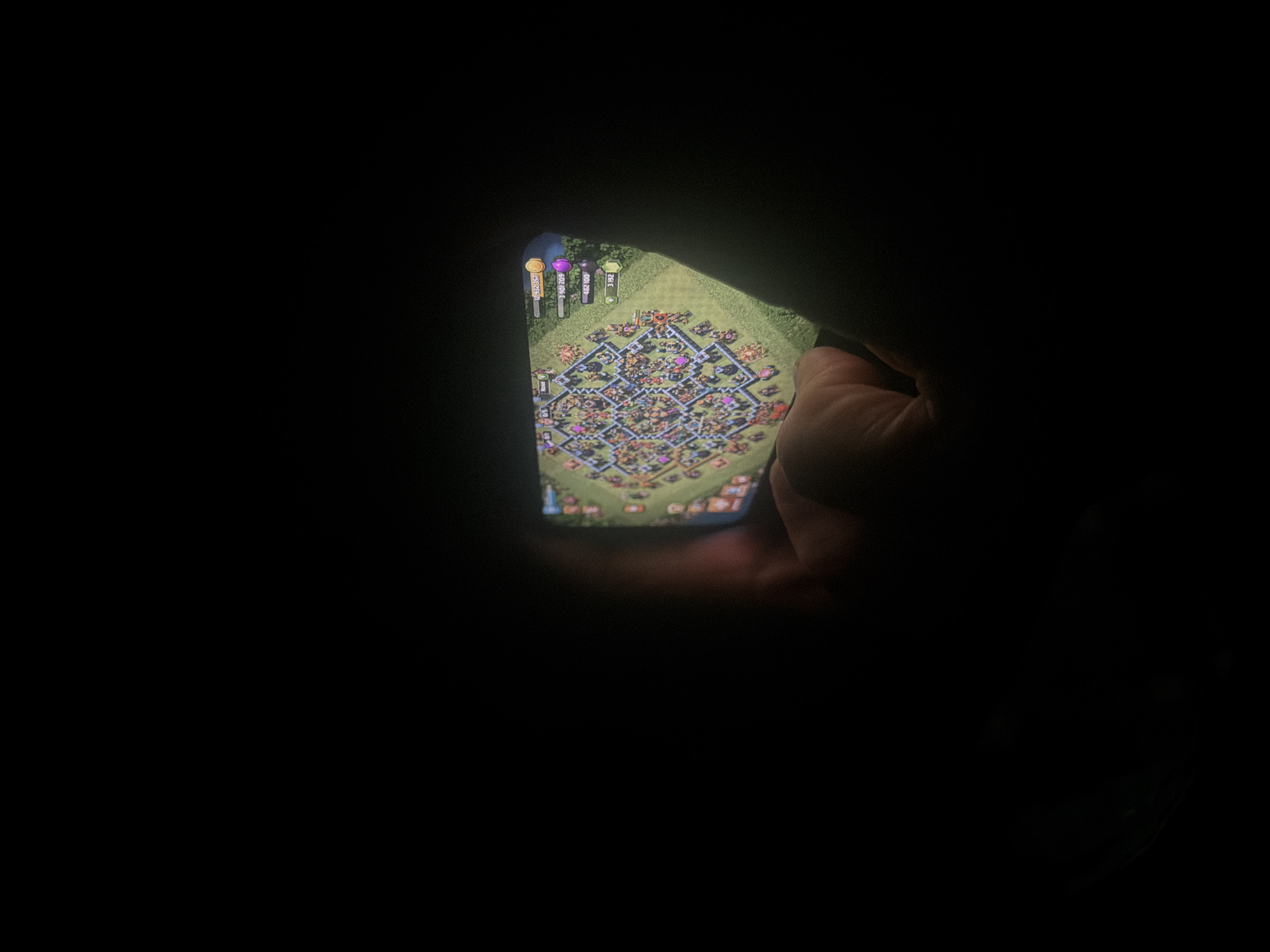The image features a person's hand holding a smartphone in a dark environment, with the phone's screen being the primary light source. The smartphone displays the mobile game "Clash of Clans," showcasing a player's village layout.
On the top-left of the phone screen, in-game resource counters are visible. These include gold, displaying "12,932,357"; elixir, showing "6,420,084"; dark elixir, at "7,162"; and another resource with a green icon, indicating "3,752." The village itself is elaborate, featuring numerous high-level buildings, defenses, and walls arranged in a structured layout. The distinct graphics confirm the game as Clash of Clans.
The background is entirely dark, suggesting the photograph was taken in a very dim setting, possibly at night or in a room with no other lights. The hand holding the phone is visible on the right side of the screen. No other objects, people, or specific location cues are discernible in the scene.