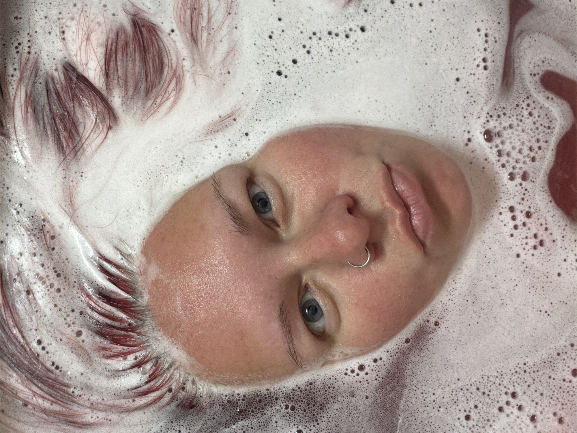 A fair-skinned person with dyed reddish-brown hair is partially submerged in a tub of bubbly, milky liquid, possibly soapy water or a bath infused with color. The person's face is visible above the waterline, tilted slightly, with clear blue eyes. A silver ring pierces their left nostril. The bubble-filled liquid surrounds their face and hair, creating a textural contrast. The color of the liquid appears to be light pink or a diluted red in certain areas. The location cannot be determined from the image alone, as it is an indoor scene. The city of Chaikovsky, Russia, cannot be confirmed as the location without additional information.