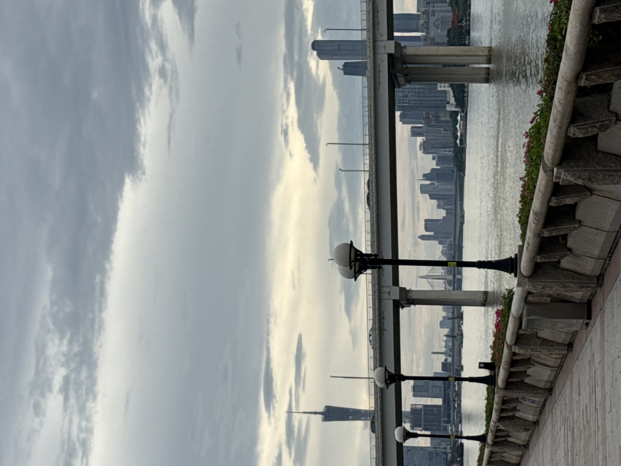 This is a photograph taken from a waterfront promenade, likely in Tianhe District, China, overlooking a wide river. In the foreground, a paved walkway runs along the left side of the image, bordered by a stone wall with a decorative pattern. Several lampposts with spherical globes are positioned along the promenade. Behind the wall, a row of green bushes with scattered pink flowers is visible.
The middle ground is dominated by a large concrete bridge spanning the river, with vehicles traveling on its surface. The bridge is supported by substantial concrete pillars. Beyond the bridge, a dense cityscape unfolds with numerous tall buildings of varying architectural styles, including skyscrapers. A prominent landmark tower, resembling the Canton Tower, is visible in the distance on the left side of the cityscape. The river itself appears calm, reflecting the overcast sky and the surrounding structures.
The sky is filled with heavy, gray clouds, suggesting a cloudy or possibly rainy day. The overall lighting is muted and diffused, casting a soft light over the scene. No people are explicitly visible in the image. The atmosphere is one of urban development and infrastructure, with a focus on the bridge and the city skyline.