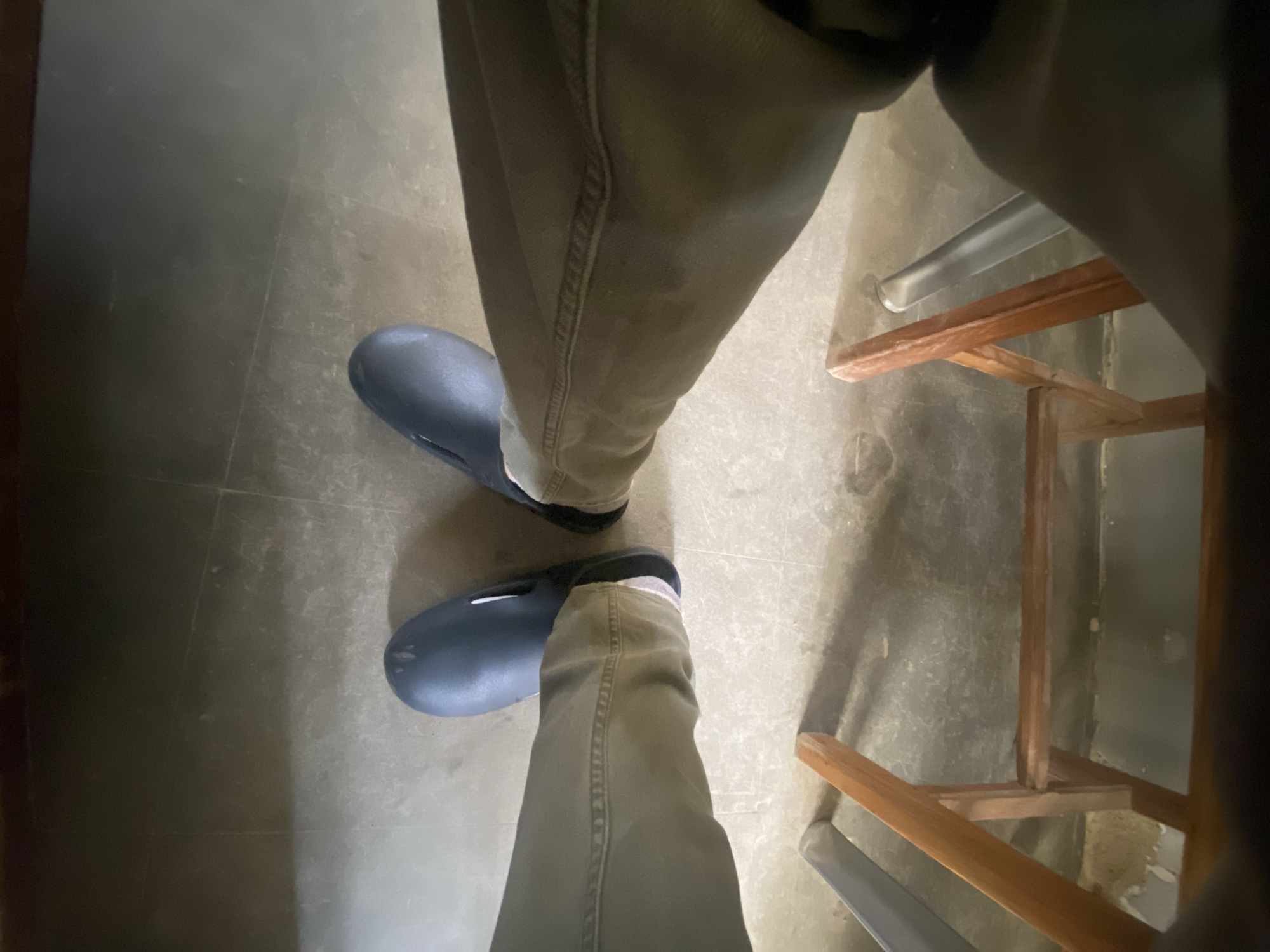 The image shows a first-person perspective of a person sitting, likely on a chair, with their legs extended forward. The individual is wearing dark blue clogs and khaki pants. The floor appears to be tiled and shows some wear and tear. In the foreground, a wooden structure, possibly part of a chair or a small stool, is visible. The lighting is dim, creating shadows. There is no visible indication of the specific location in the city of Jos, Nigeria, beyond what can be inferred from the general appearance of the indoor environment.