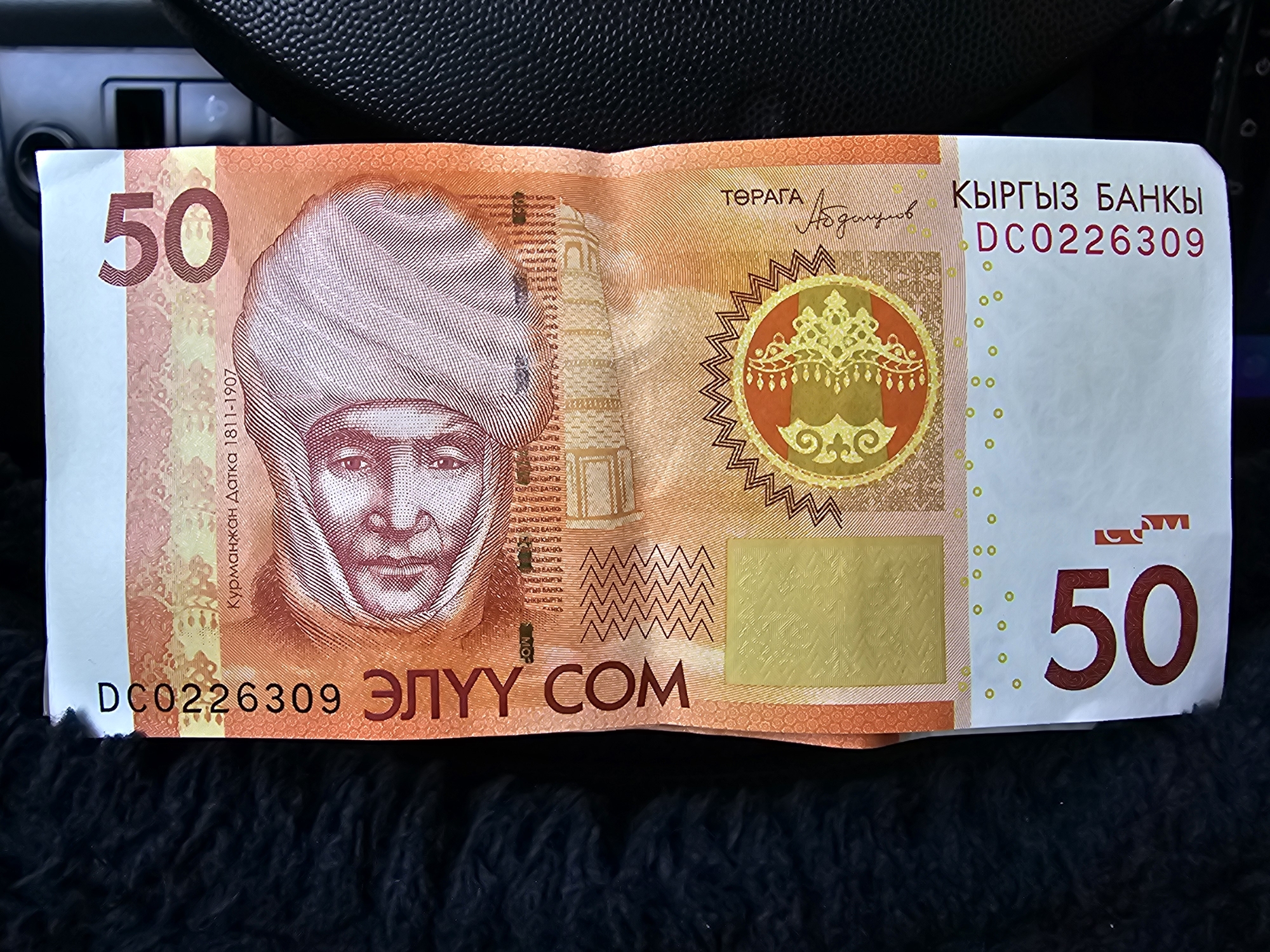 The image shows a close-up of a Kyrgyzstan 50 som banknote. The banknote is laid out horizontally and appears to be on a dark, textured surface. The front of the banknote features a portrait of a man wearing a traditional Kyrgyz hat and a detailed background with architectural elements and inscriptions. The denomination "50" is prominently displayed in large red numerals on both the left and right sides of the banknote. Text in the Kyrgyz language is visible, including the name of the bank and other identifying marks. The banknote is creased and slightly worn, suggesting it has been in circulation. The background of the image is out of focus, but appears to be part of a car's interior, with a black, textured surface that could be a dashboard or seat. The lighting suggests it is indoors or in a shaded area. No specific location in the city of Özgön, Kyrgyzstan, can be determined from this image.