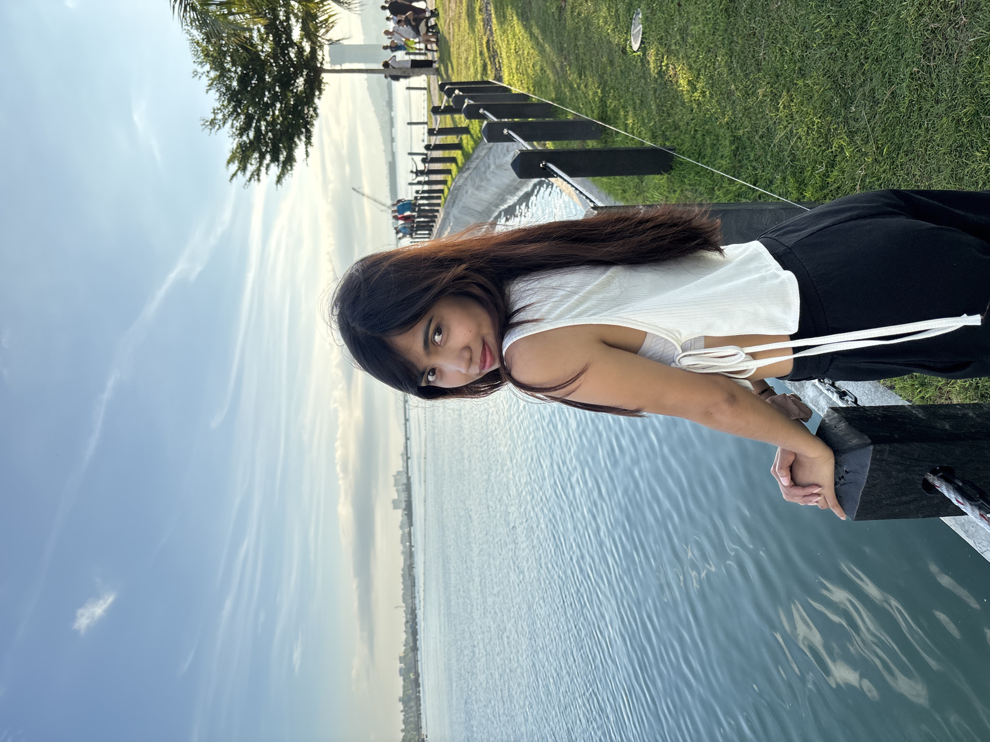 A young woman with long, dark hair is positioned in the foreground, looking over her shoulder towards the camera. She is wearing a white, sleeveless crop top with tie accents and black pants. She stands near a black fence bordering a body of water. The water, which appears to be a bay or strait, exhibits gentle ripples and reflects the sky. Beyond the fence, a grassy area slopes upwards, with a paved pathway and more black fencing. Several individuals are visible in the background, walking and sitting along the pathway. A tree with lush green foliage stands to the right of the frame, casting shadows on the grass. In the distance, a city skyline with buildings and a tall tower can be seen across the water, under a bright blue sky with scattered white clouds. The scene suggests a public park or waterfront promenade in Davao City, Philippines, likely during the late afternoon or early evening due to the warm lighting and long shadows.