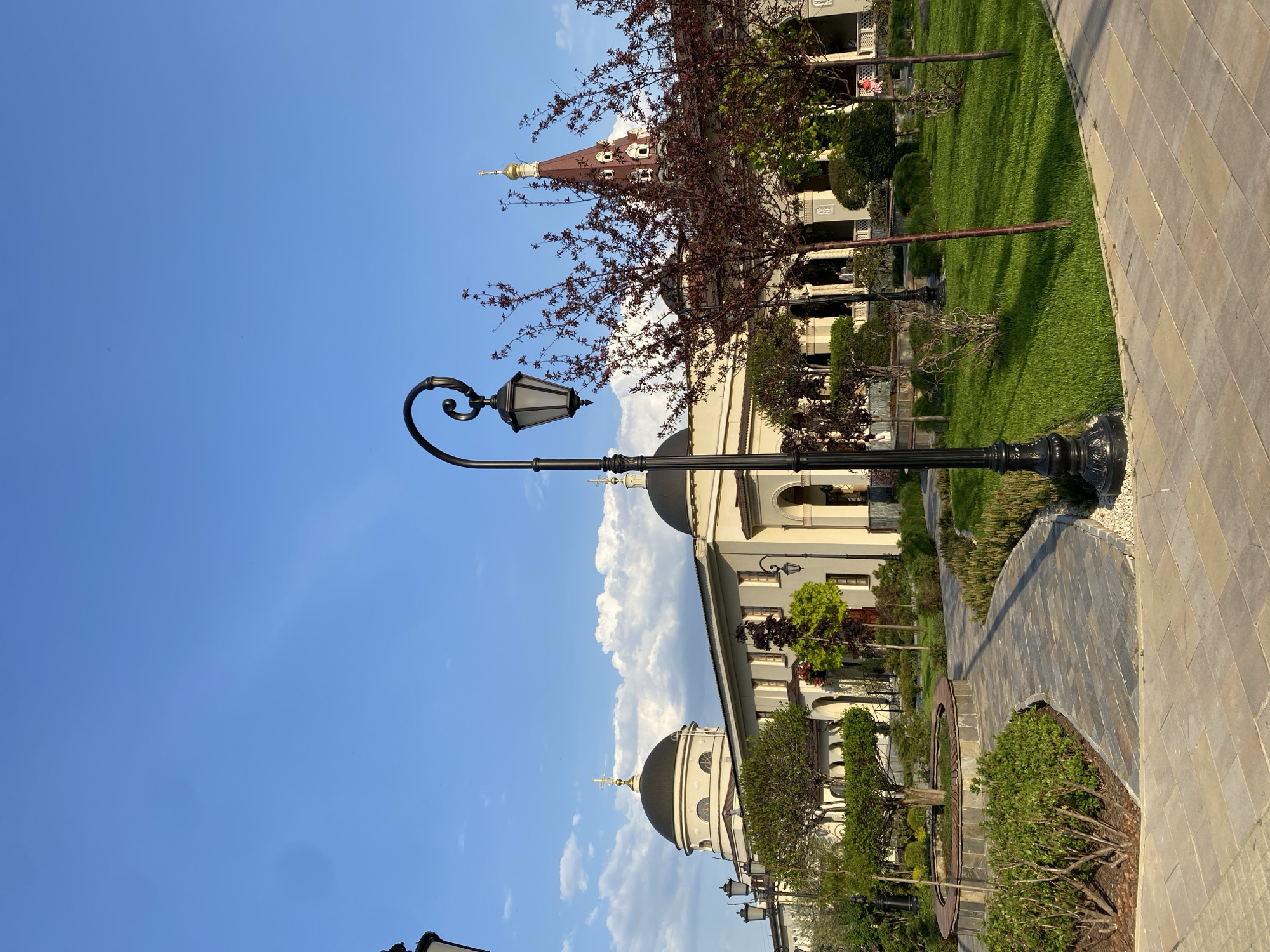 The image depicts a street scene in Yekaterinburg, Russia, featuring a black ornate lamppost in the foreground with a traditional lantern. The sky is mostly clear blue with scattered white clouds. Behind the lamppost is a white building with black domes topped with golden crosses, suggesting a church or cathedral. Part of a red tower is visible in the background. A sidewalk and landscaped areas with green grass and bushes are visible to the right of the building. There are no people visible.