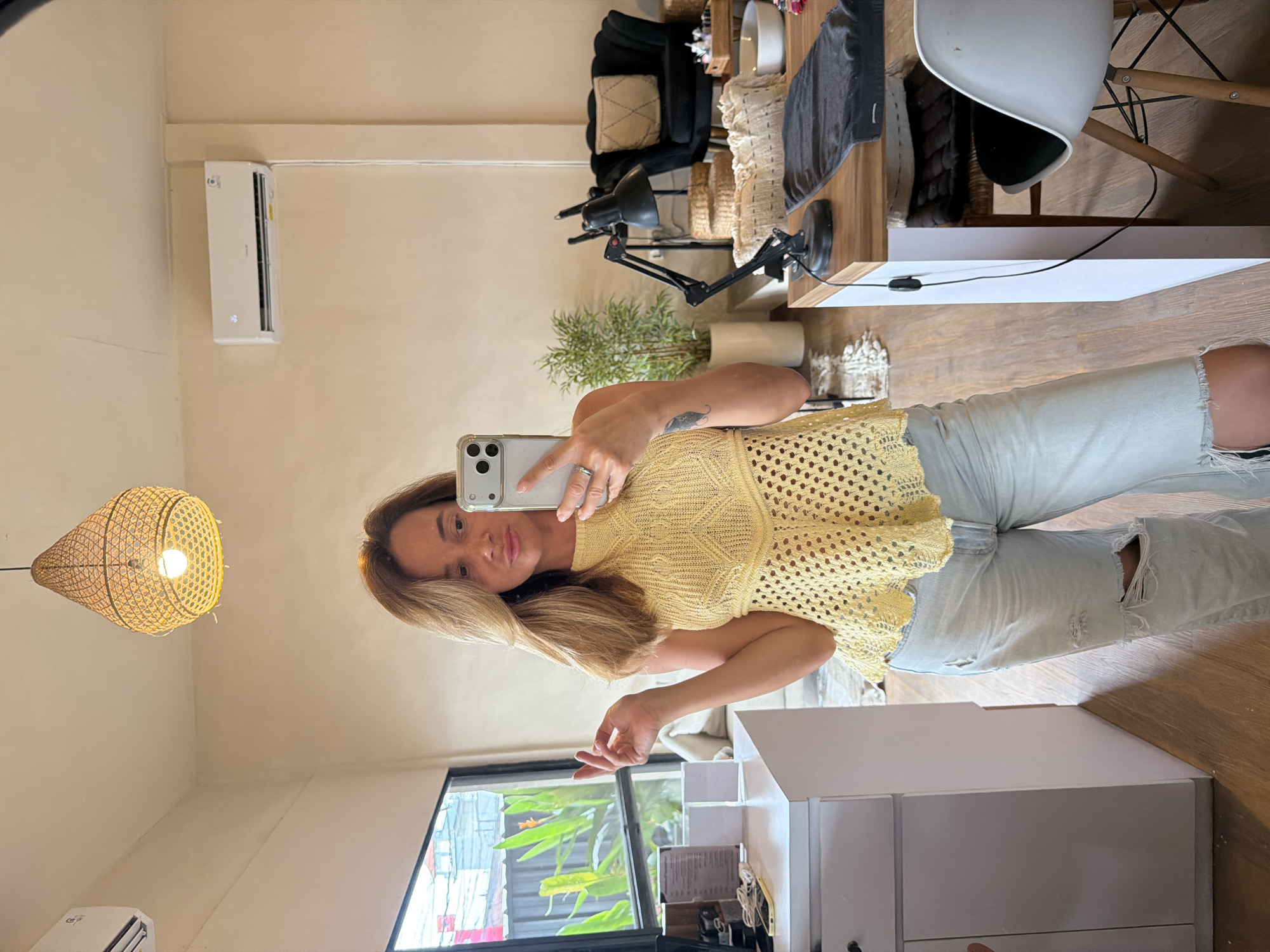 A woman with shoulder-length blonde hair takes a mirror selfie in a room. She is wearing a yellow crochet halter top and light wash ripped jeans. The room has cream-colored walls, a wooden floor, and a large window on the left showing greenery outside. To the right, there is a desk with a black lamp, a black armchair, and wicker baskets. An air conditioning unit is mounted high on the wall. The location is in North Kuta, Indonesia.