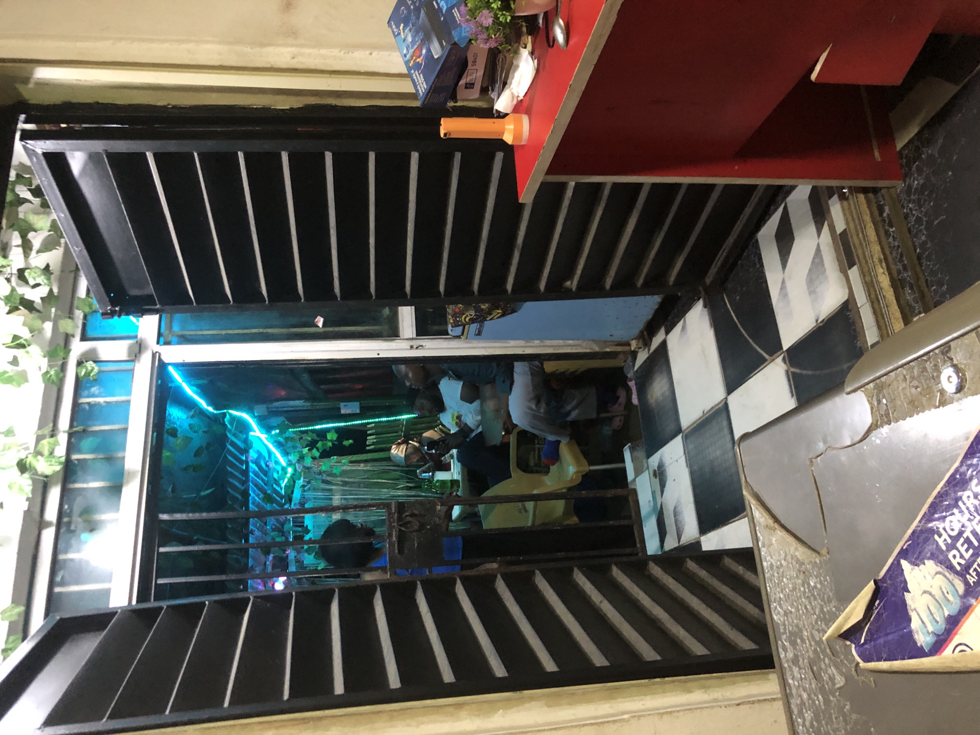 The image depicts the entrance to a dimly lit establishment in Ado, Nigeria. The foreground shows a damaged metallic surface with a partial view of a purple and white sticker. Beyond this, a black metal gate, partially ajar, reveals the interior. Through the gate, several individuals are seated at tables in a room illuminated by neon blue and green lights, with artificial vines hanging from the ceiling. To the right of the entrance, a red counter is visible, holding a bright orange flashlight, a box labeled "DStv," and a small potted plant. The floor inside the establishment features a black and white checkered pattern. The overall impression is of a simple, perhaps informal, business or social gathering space.