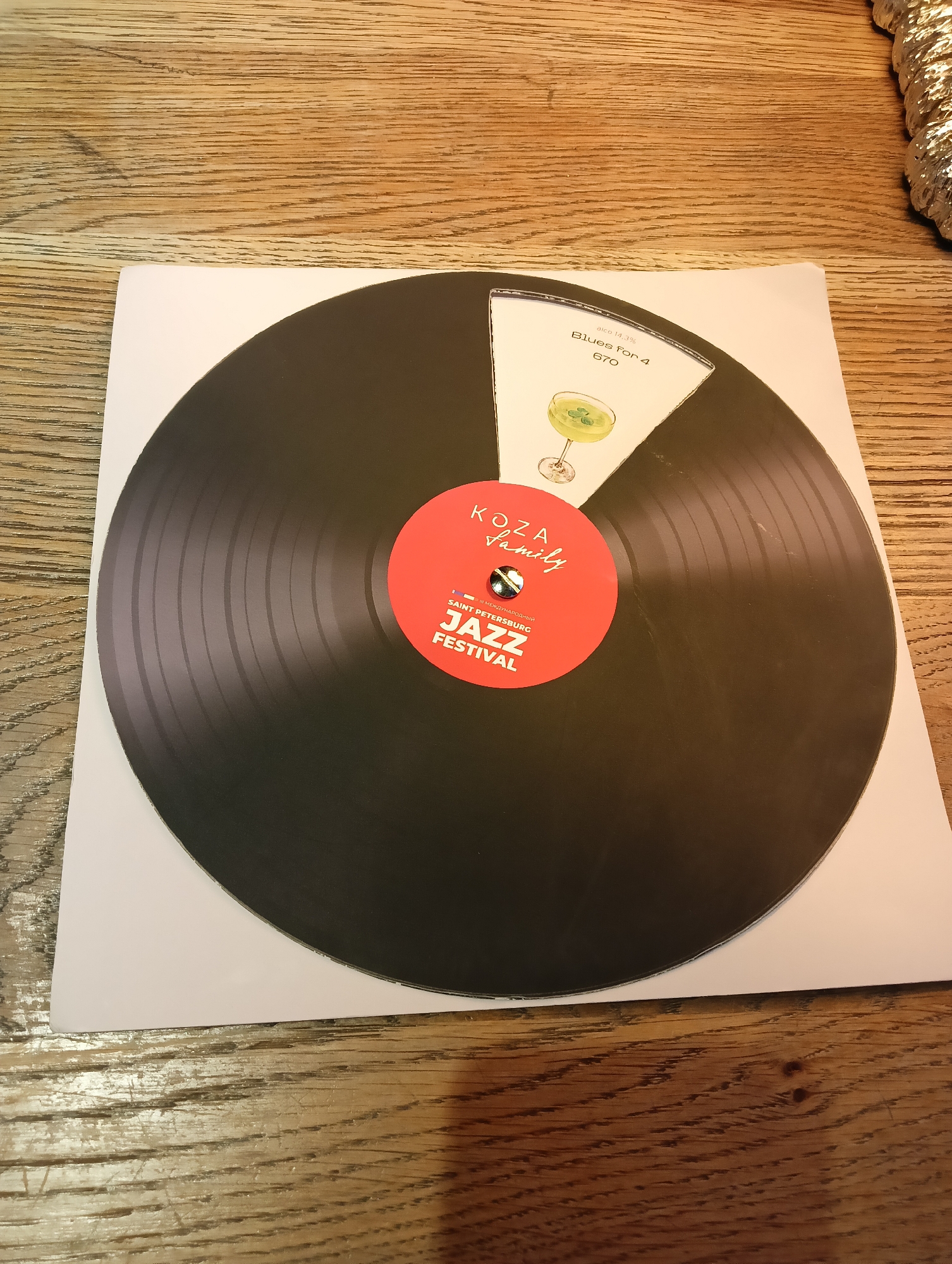 A close-up overhead shot shows a black vinyl record-shaped graphic placed on a white sheet of paper, which rests on a wooden table. The record graphic features a red label in the center with text that reads "KOZA Family" and "SAINT PETERSBURG JAZZ FESTIVAL". A section of the record is a wedge-shaped cut-out revealing a light-colored background with an illustration of a cocktail glass and text that includes "alco 11,3%", "Blues For 4", and "670". The wooden table surface exhibits a distressed, textured grain pattern with visible knots and cracks. The scene is evenly lit with no shadows or prominent light sources visible. No people are present in the image.