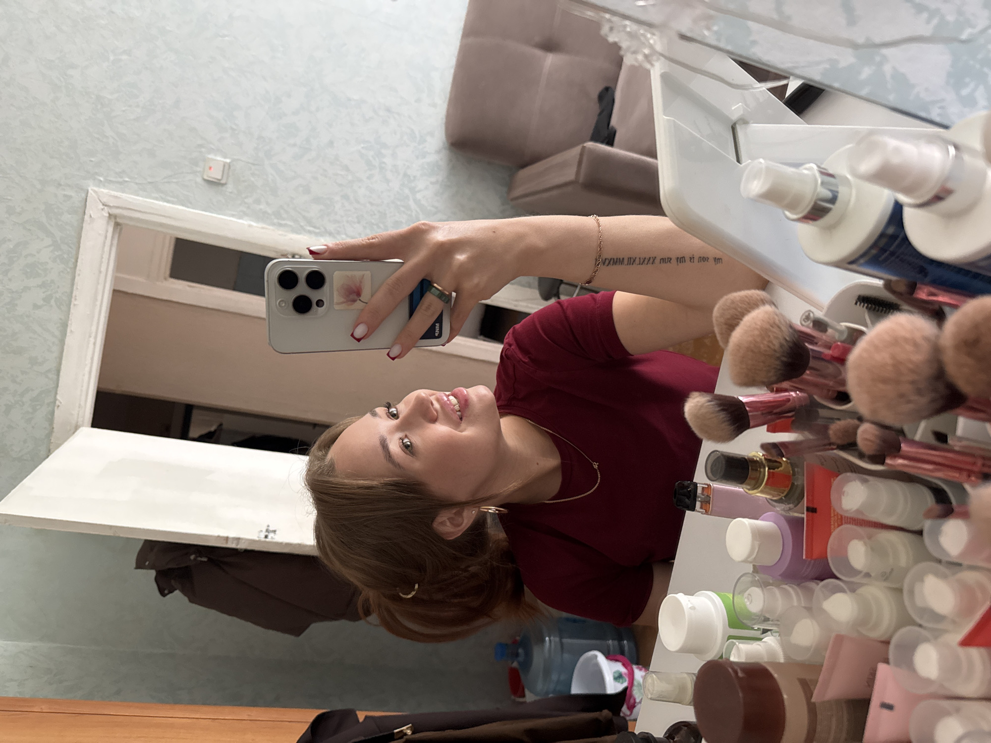 A young woman, likely in her late teens or early twenties, is taking a selfie in a mirror. She has light brown hair, parted in the middle, and is wearing a dark red t-shirt. Her left hand, adorned with a green ring and a rose-themed phone case on her phone, is visible as she holds up the device. A tattoo of Roman numerals and text is visible on her forearm. She is smiling and looking up and to her right.
The background suggests an indoor setting, possibly a dressing area or vanity. A mirror is the primary object reflecting the scene. To the right of the mirror, a collection of makeup brushes and various cosmetic bottles are neatly arranged in a white organizer. Some of the bottles have white or clear caps and appear to contain lotions or sprays. In the reflection of the mirror, a portion of a light blue wall with a textured pattern can be seen, along with a white doorway and a glimpse of furniture, possibly a sofa, in a neutral tone. A small, square white object with a red light is mounted on the wall. The overall lighting suggests it is daytime, likely indoors. The mood appears relaxed and casual. No specific weather conditions are discernible. The location context of Chaikovsky, Russia, is provided but not explicitly visible in the image itself.