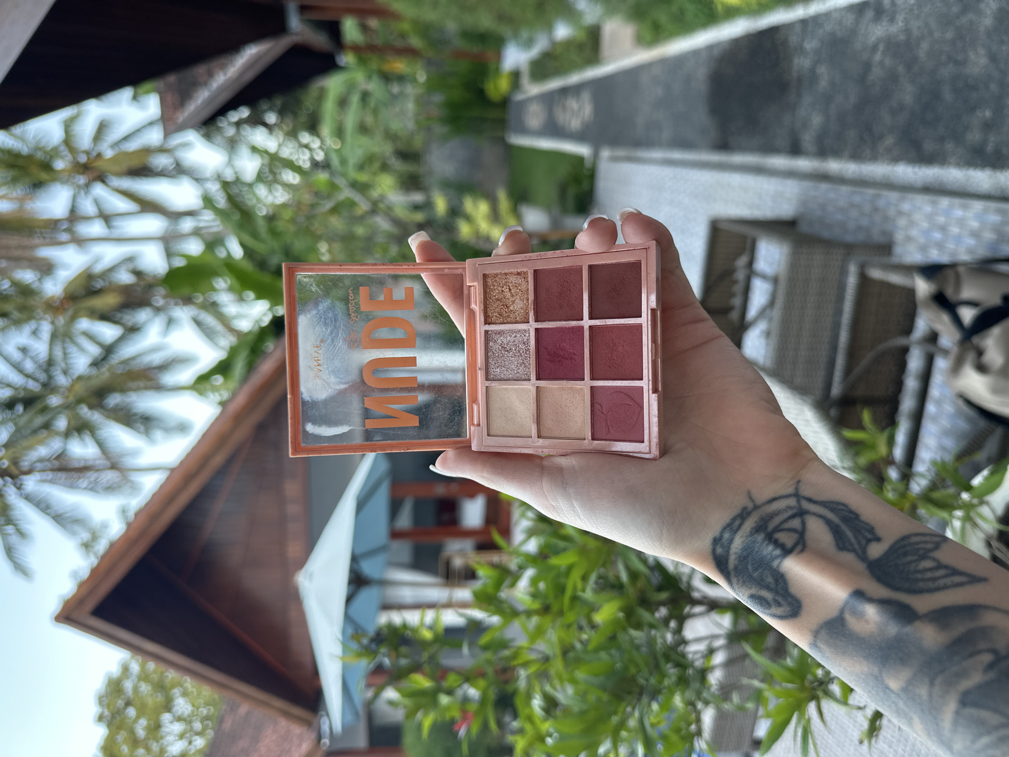 A woman's left hand holds an open "NUDE" eyeshadow palette in an outdoor setting. The palette has nine shades in varying neutral and reddish tones. The woman has a tattoo on her forearm. The background includes a building with a peaked roof, greenery, and a tiled walkway with seating. The location is Nusa Penida, Indonesia, as indicated by the building's architecture and tropical vegetation.