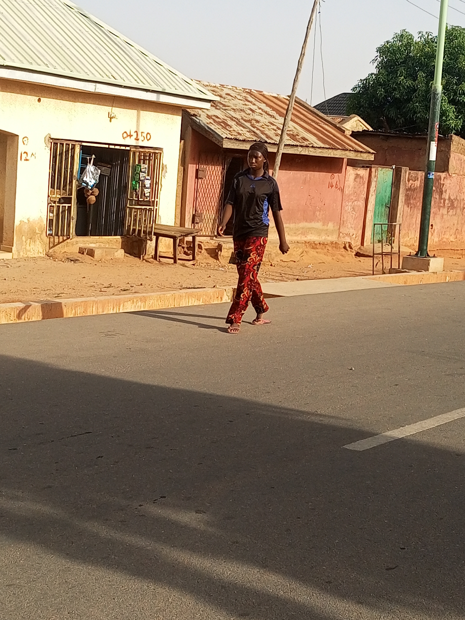 The photograph captures a woman walking along a paved road in what appears to be a residential area of Tempure, Nigeria. The woman is wearing a dark-colored t-shirt with blue accents and patterned red and black pants. She is walking barefoot.
The environment consists of buildings with varying architectural styles. One building has a corrugated metal roof, and another appears to be a small store with barred windows. There's a wooden bench outside the store. The buildings are painted in faded shades of peach and beige. A green metal gate or door and a concrete post with green paint are visible near a wall. The road is shaded, and a white line marking is visible on the road surface. A utility pole stands in the background.