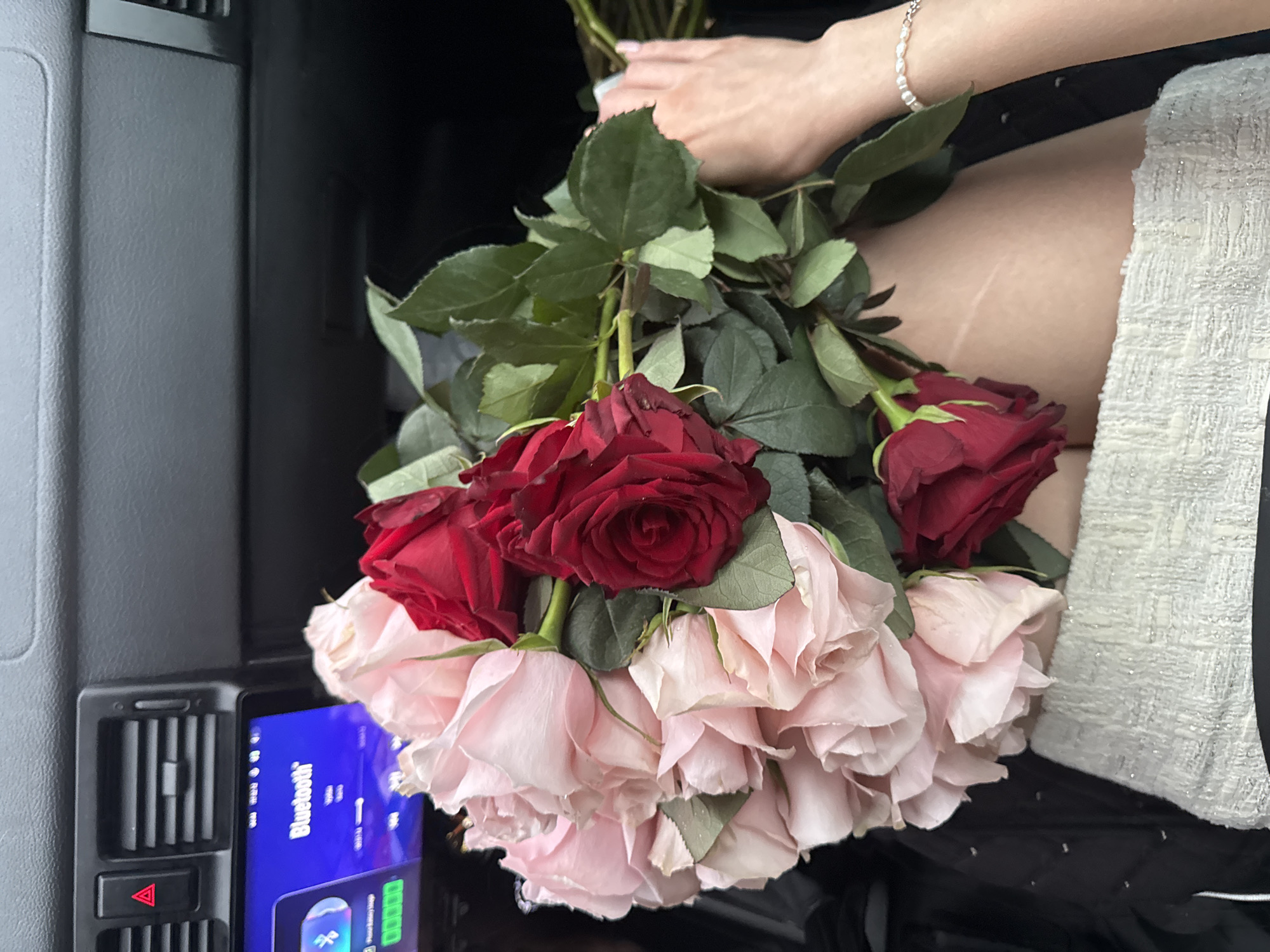 A bouquet of light pink and dark red roses is held by a person in a car. The bouquet is positioned in the foreground, with the stems partially obscured by the person's legs and clothing. The person is wearing a white textured skirt and a dark jacket. A bracelet with small pearls is visible on their wrist. In the background, the interior of a car is visible, including a car stereo with a Bluetooth interface, a hazard light button, and air vents. The car appears to be parked, with the lighting suggesting it is daytime. The specific location of the city of Perm, Russia, is not discernible from the image.