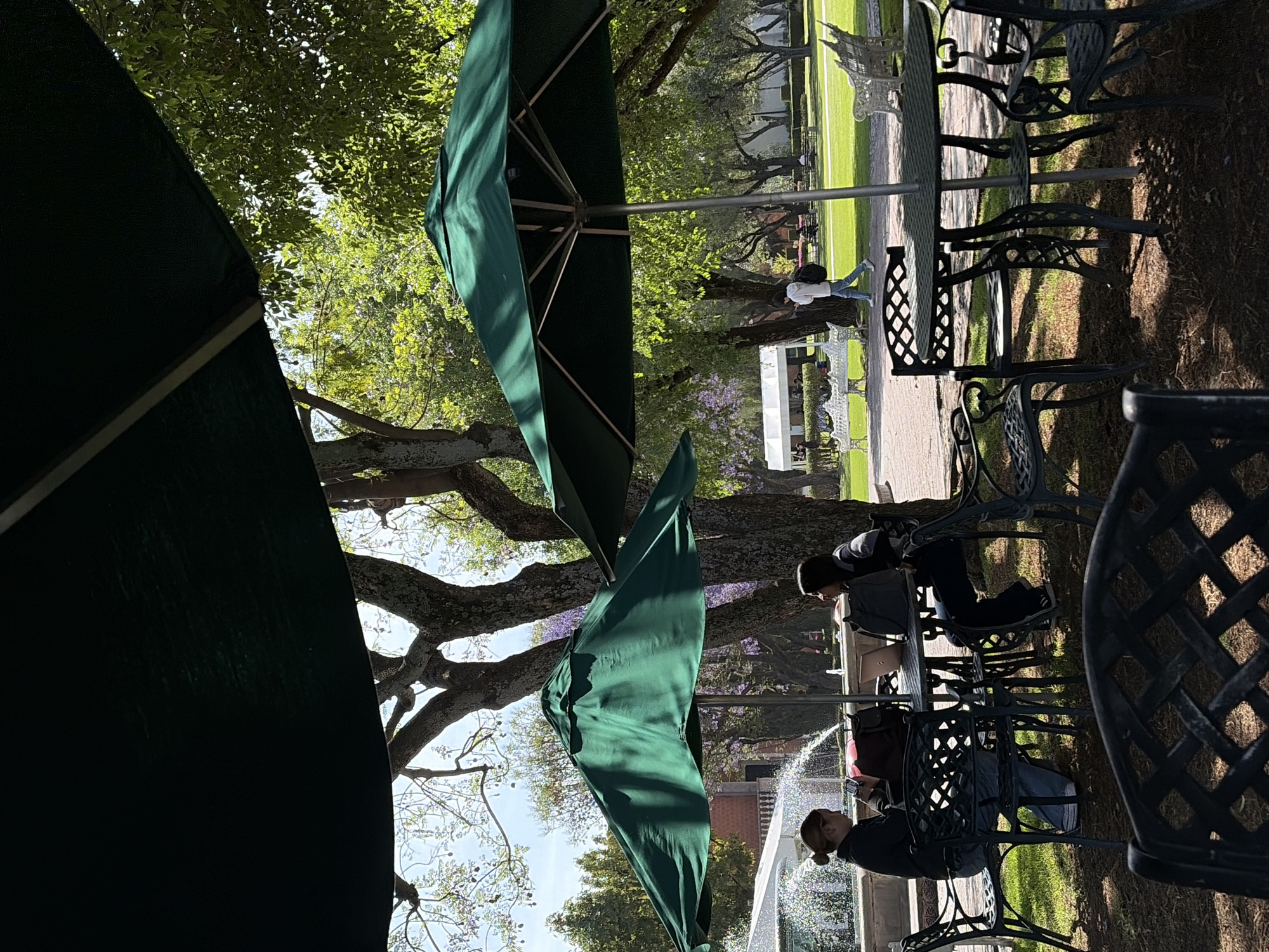 The image shows an outdoor café setting in San Andrés Cholula, Mexico, with people seated at tables under green umbrellas. The café appears to be in a park or garden, with trees visible in the background. The foreground shows wrought iron chairs. A fountain with spraying water can be seen in the distance. People are sitting at the tables, with one person seemingly taking a photo. The scene is brightly lit, suggesting it is daytime.
