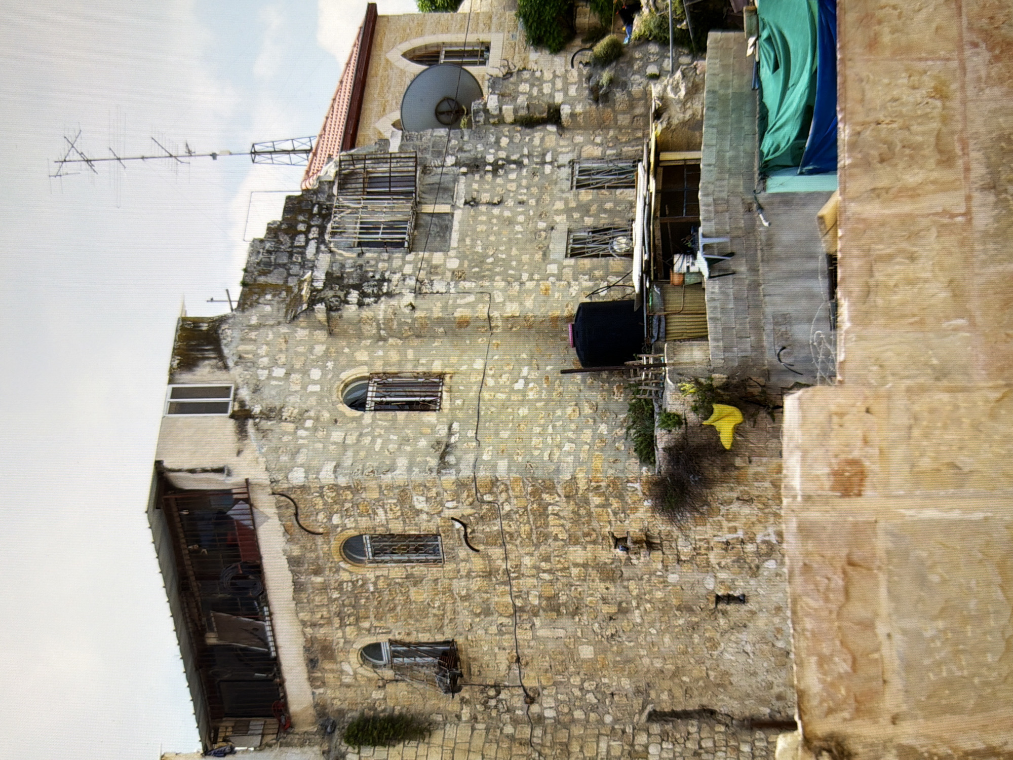 The image shows a stone building with multiple stories and windows. The building appears old and weathered, with some areas showing signs of disrepair. There are several objects visible on and around the building, including a satellite dish, an antenna, a water tank, and some tarps. The surrounding environment appears to be an urban area with other buildings and vegetation visible. The scene is set in the city of Kyiv, Ukraine.