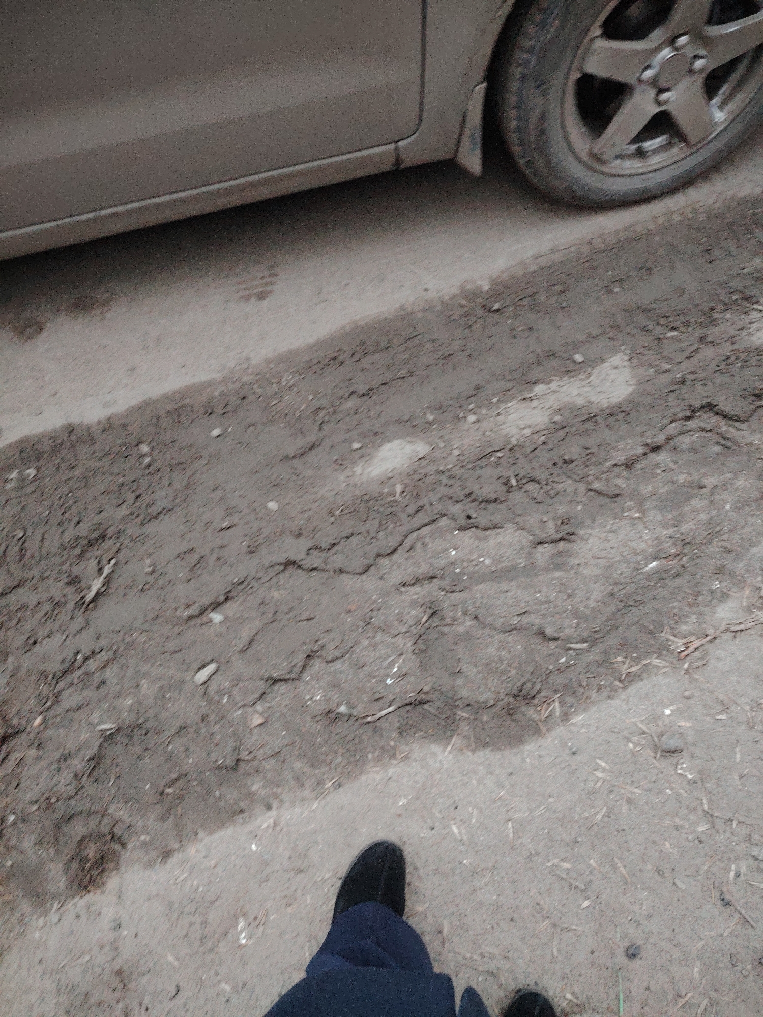 The image presents an outdoor scene. A portion of a car, including the lower body and a front tire, is visible in the upper section of the image. The car's body appears to be a neutral color. The tire is dirty, with visible wear. The surface below the car consists of cracked and uneven dirt or soil. The ground is a mix of gray and brown tones, suggesting it might be damp. In the lower section of the image, a pair of feet wearing black shoes and dark blue pants are seen. The location is unspecified, but the description requests the city of Sarapul, Russia to be included. Without additional information, the exact location in Sarapul cannot be determined.