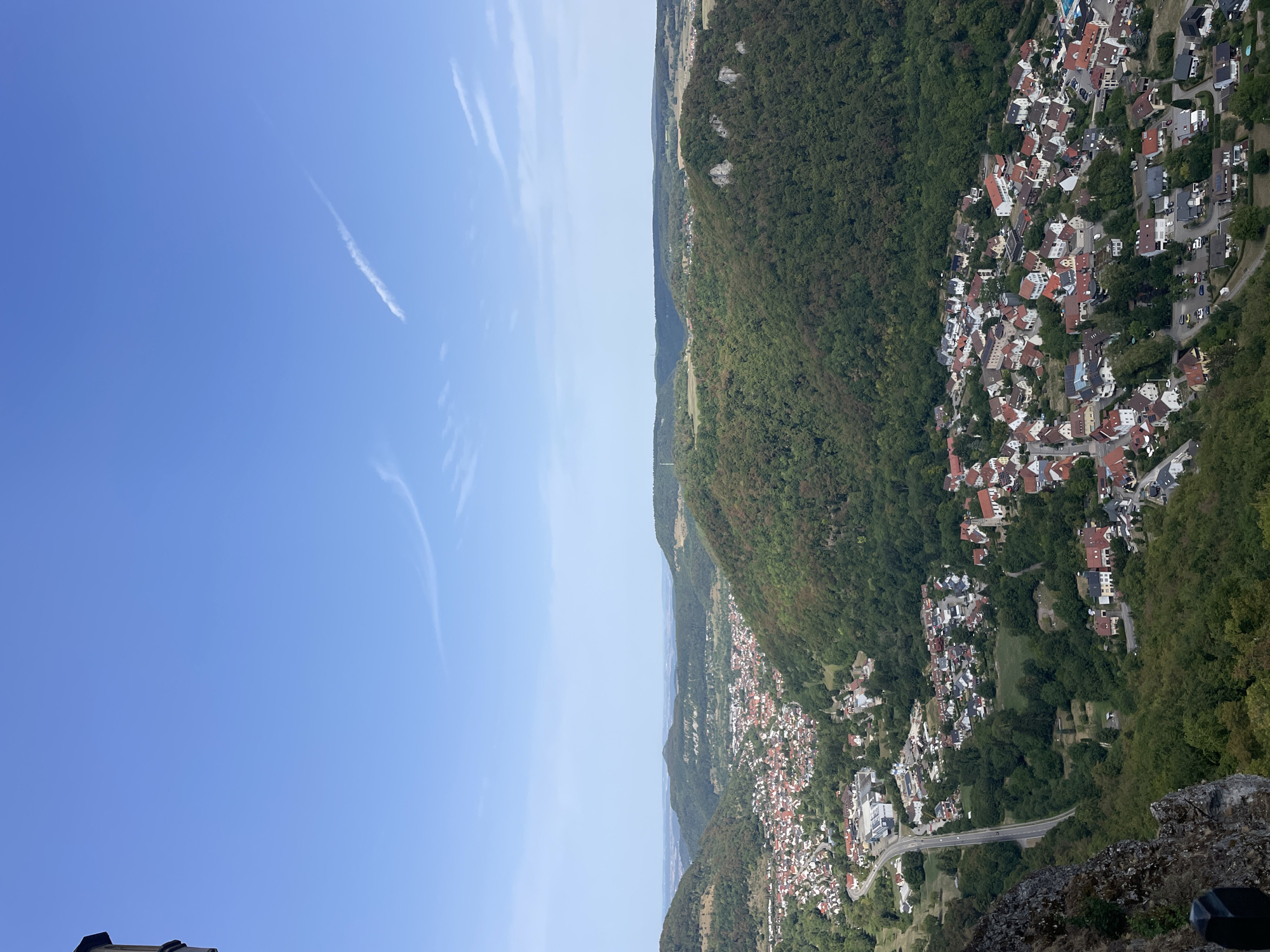 A high-angle, outdoor, daytime shot captures a panoramic view of a valley town nestled between lush green hills under a clear blue sky with wispy clouds. The town, characterized by its red-tiled roofs and dense housing, occupies the valley floor. A winding road snakes through the town, connecting various parts. The foreground features a rocky cliff face and a dark vertical post, suggesting the vantage point is from a high elevation, possibly a lookout or a castle. The background showcases rolling hills that fade into the distance, with a hint of haze. The scene is serene, with no people or animals visible. The overall impression is one of natural beauty and human settlement coexisting peacefully within a picturesque landscape.